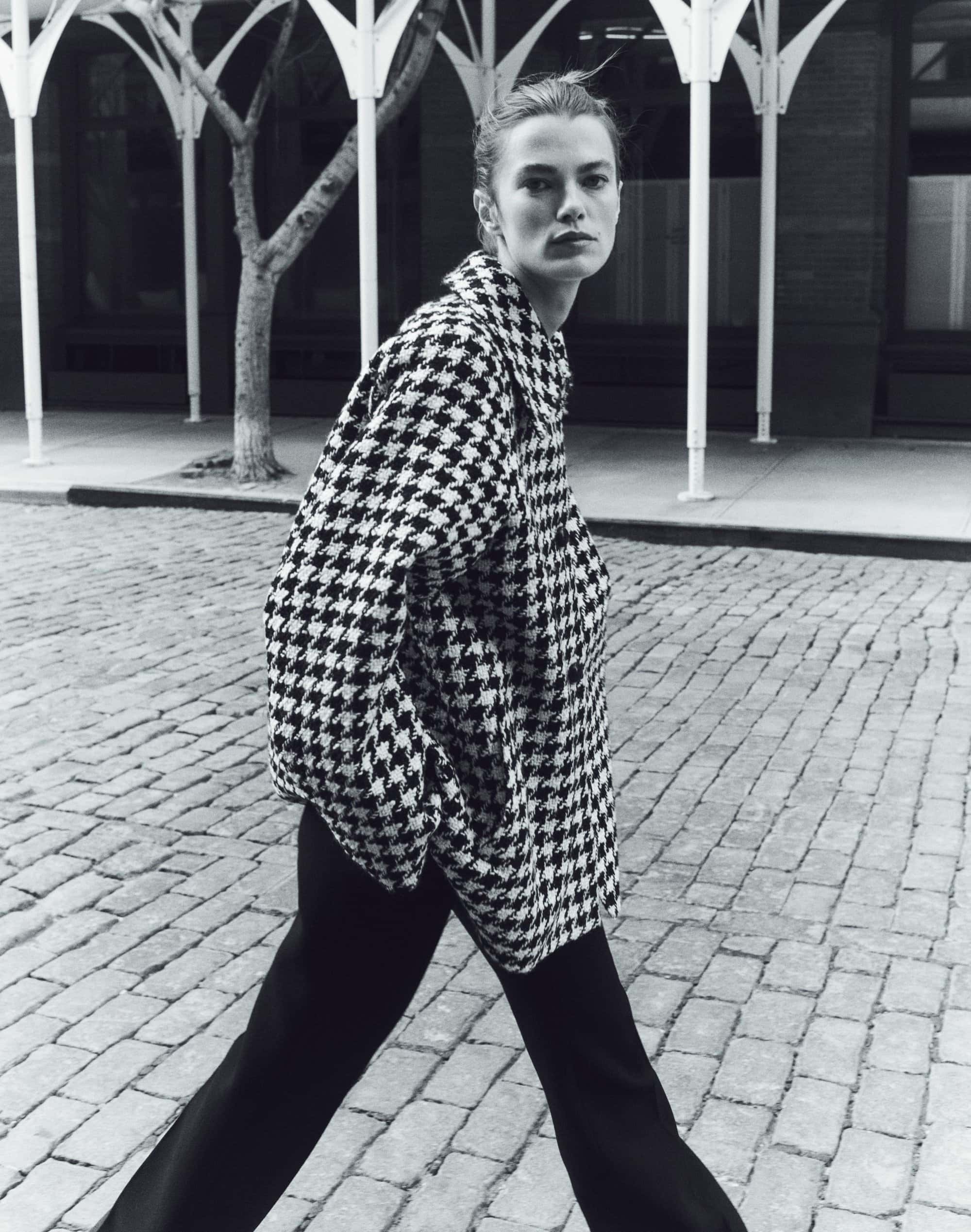 Model in houndstooth jacket and black trousers walking on a cobblestone street by white columns.