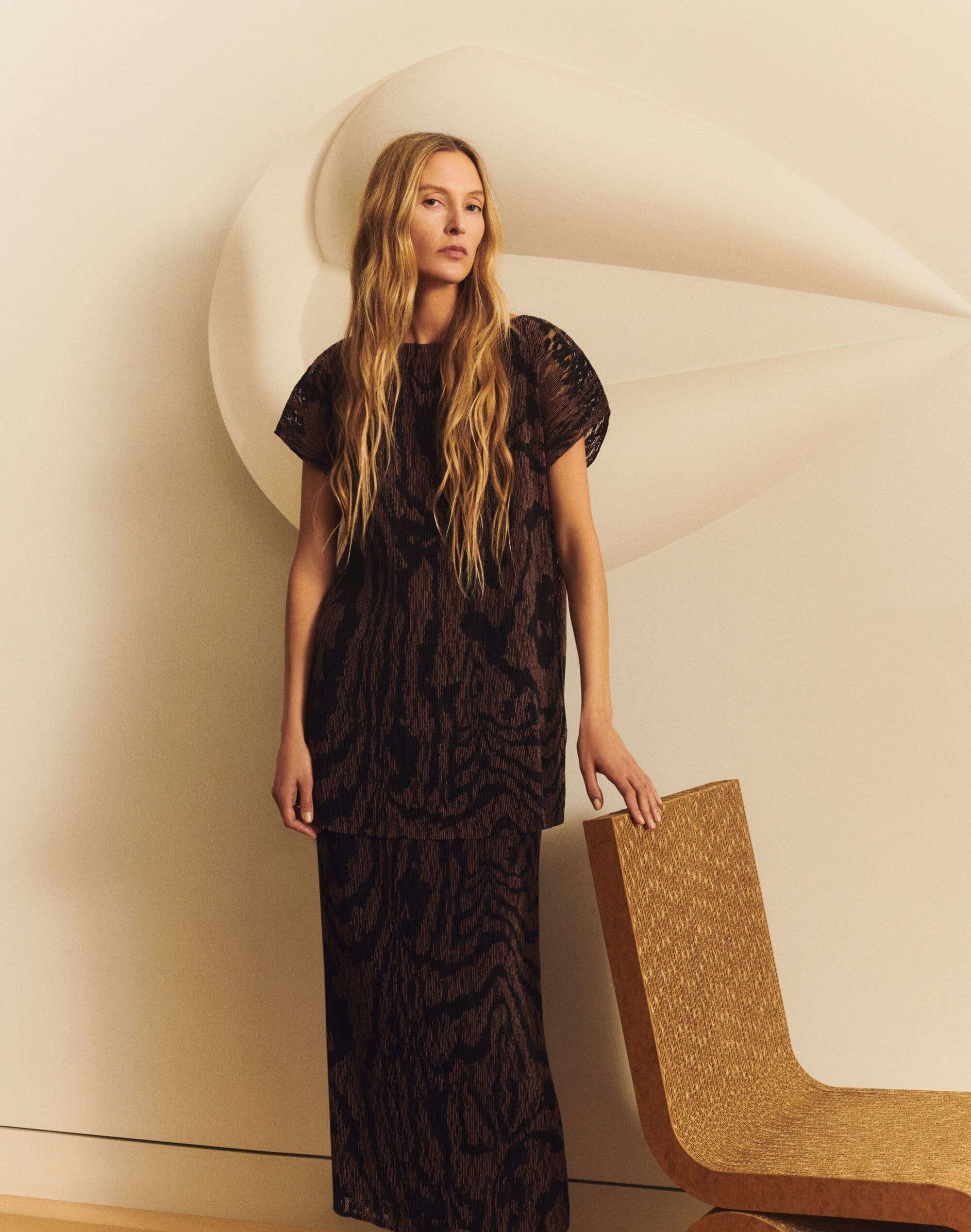 Model standing next to a textured chair wearing a dark patterned top and matching skirt in a studio setting