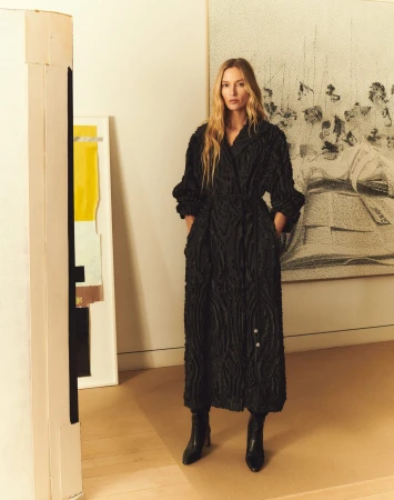 Model wearing a long black textured coat and black boots standing in an interior space with large artworks on the wall.