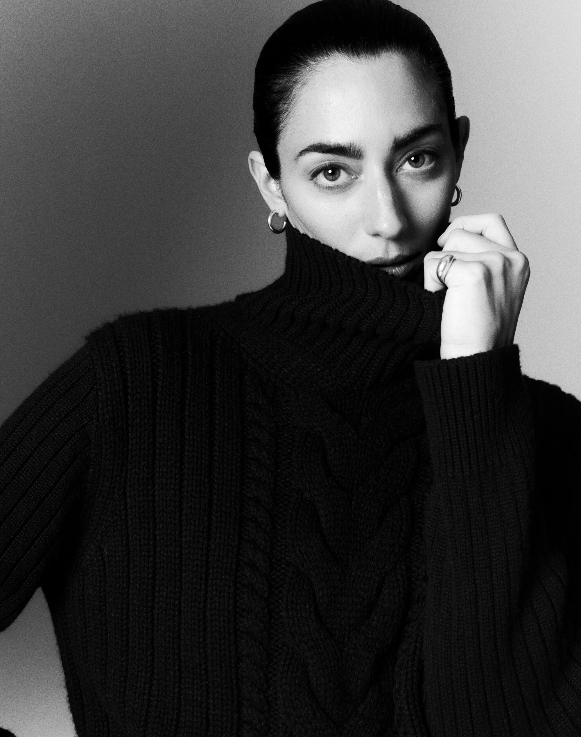 Model in black turtleneck sweater pulling the collar up near her face against a dark grey seamless studio backdrop