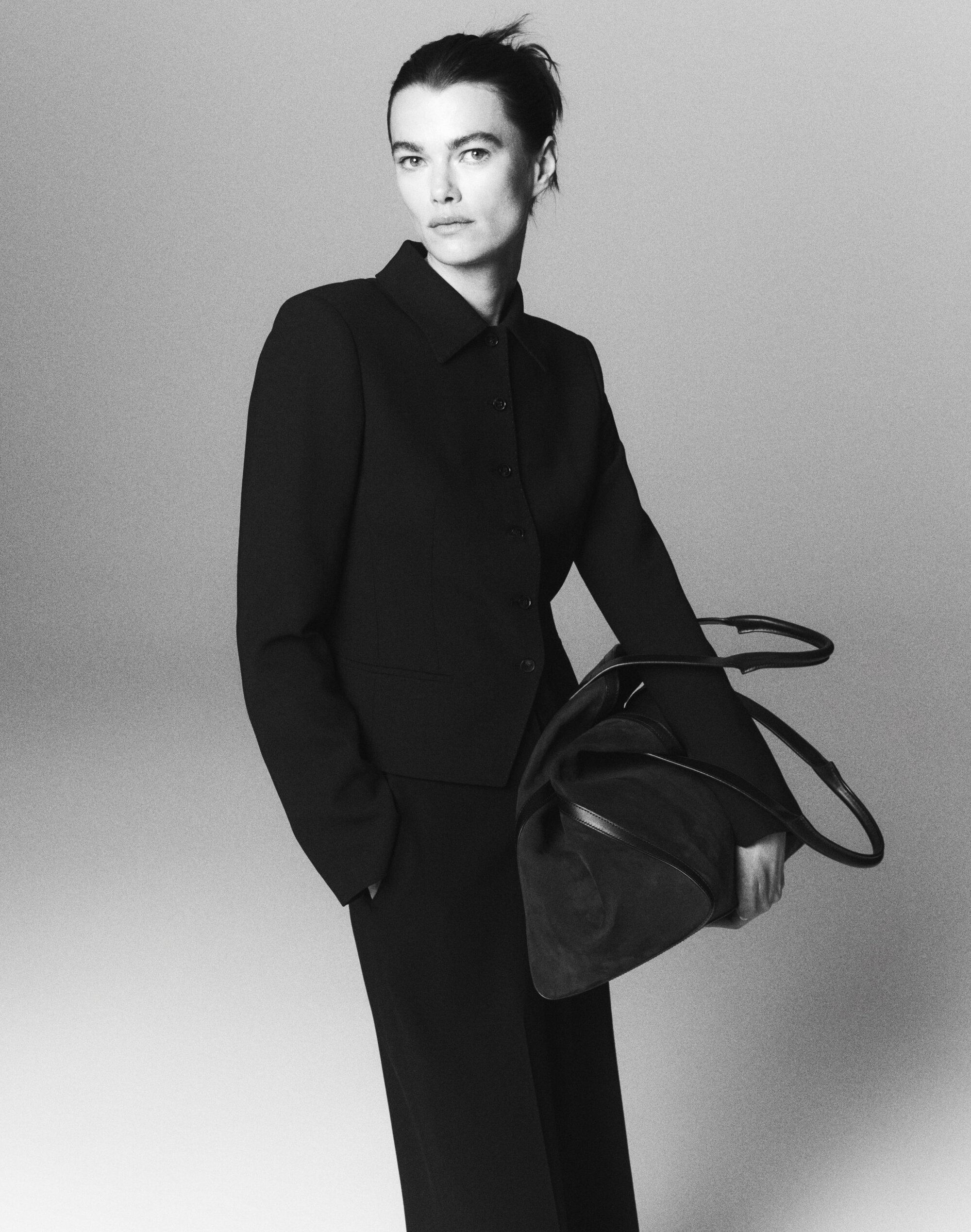 Model in black buttoned jacket holding a large dark handbag against a grey studio background