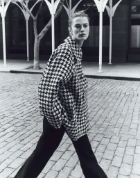 Model in houndstooth jacket and black trousers walking on a cobblestone street by white columns.