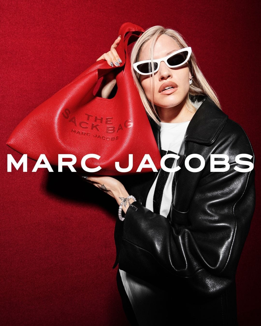 Blonde woman wearing white cat-eye sunglasses and a black leather jacket, lifting a red Marc Jacobs leather handbag against a textured red background