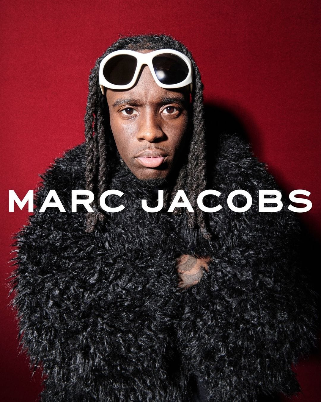 Kai Cenat wearing white sunglasses and a textured black jacket against a dark red backdrop for Marc Jacobs Fall 2024 campaign.