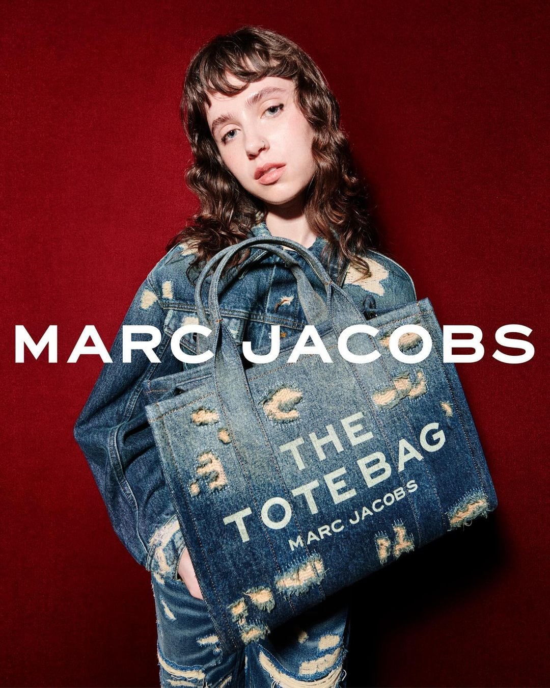 Clairo wearing a distressed denim jacket holding a denim tote bag against a dark red backdrop for Marc Jacobs Fall 2024 campaign