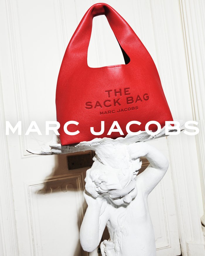 Bright red leather handbag resting atop a white carved cherub statue against a paneled door