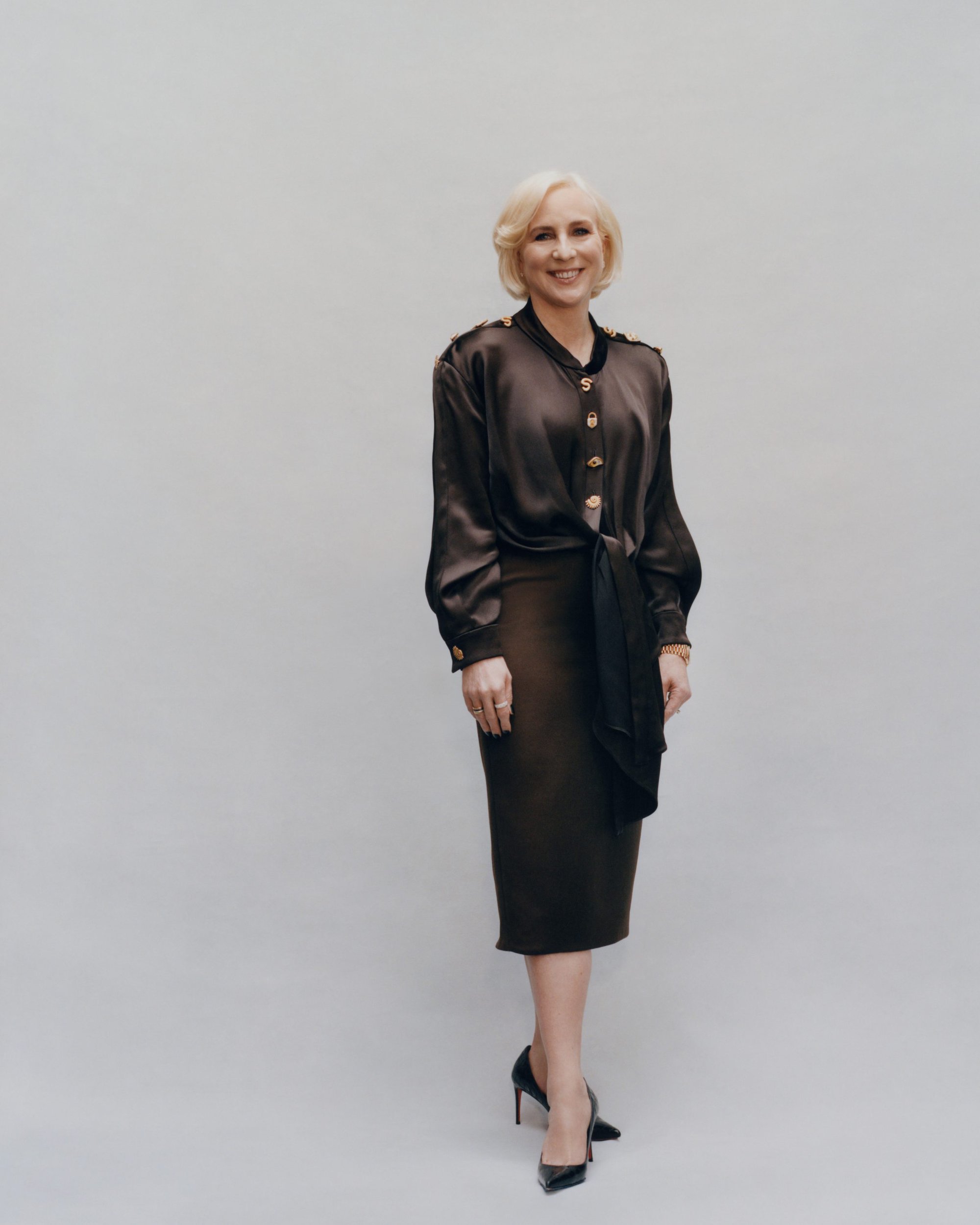 Amy Fine Collins standing against a grey seamless studio backdrop wearing a dark brown silk blouse and pencil skirt