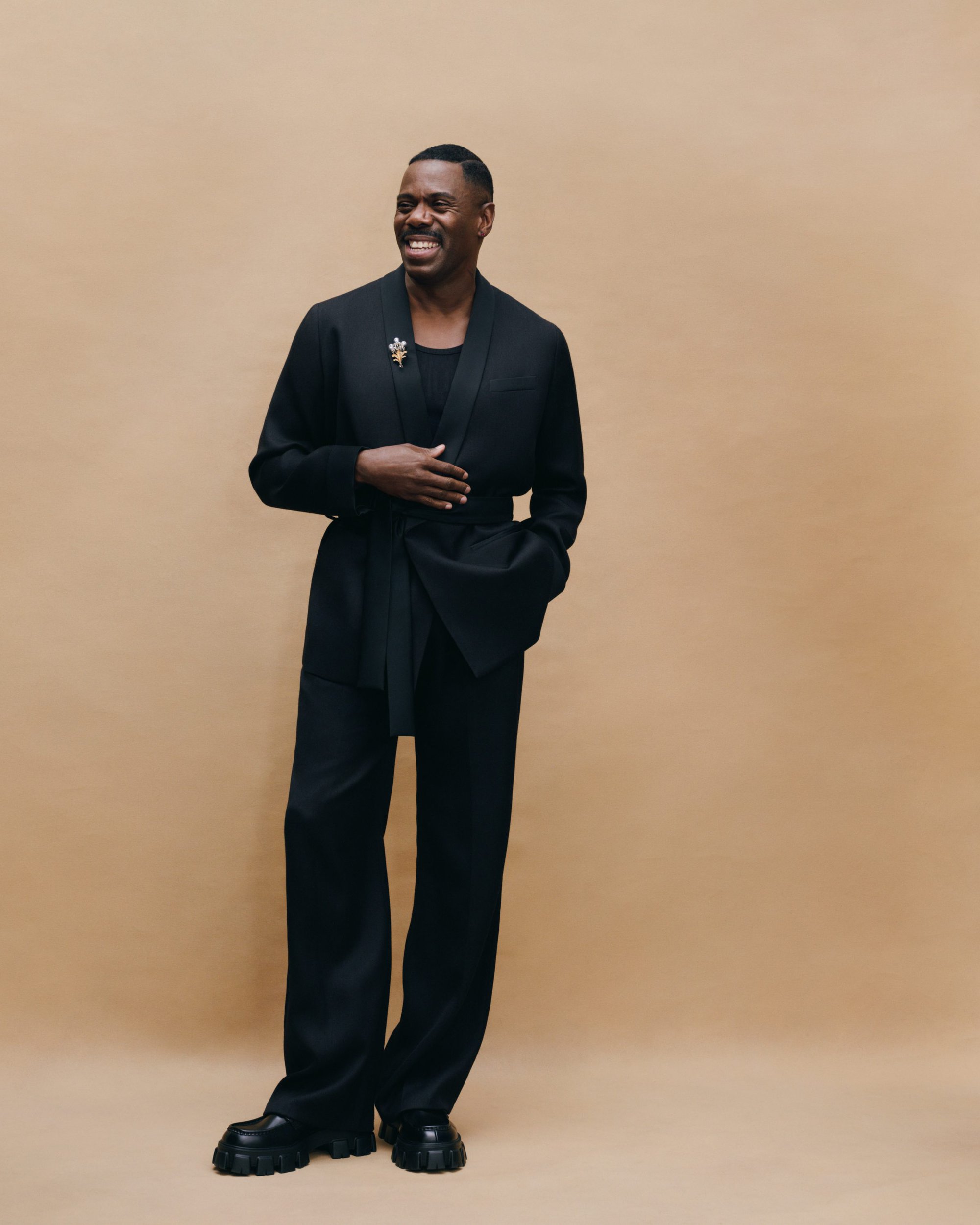 Colman Domingo wearing a black belted blazer and matching trousers against a warm studio backdrop.