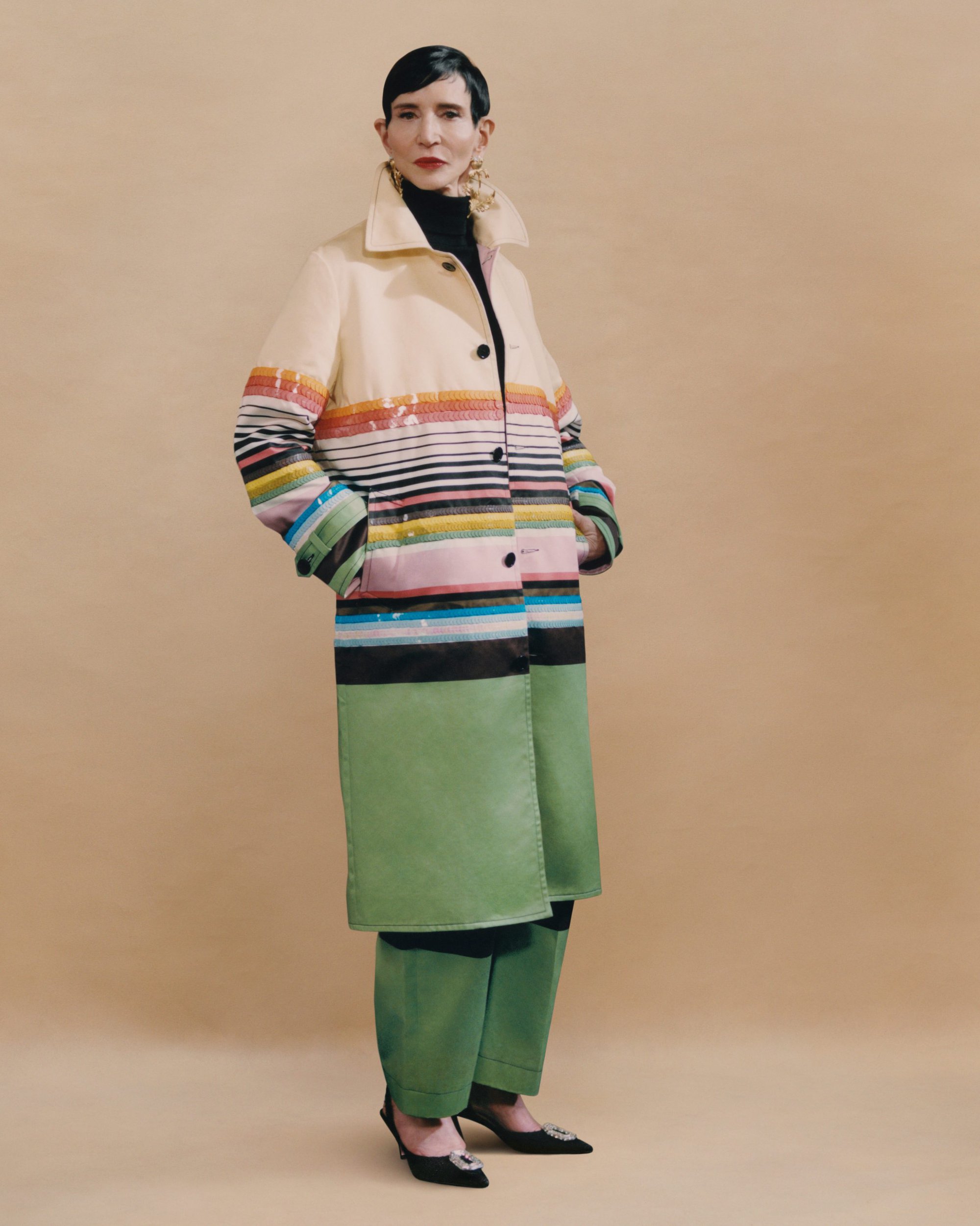 Amy Fine Collins wearing a multicolored striped coat and green trousers against a neutral studio backdrop