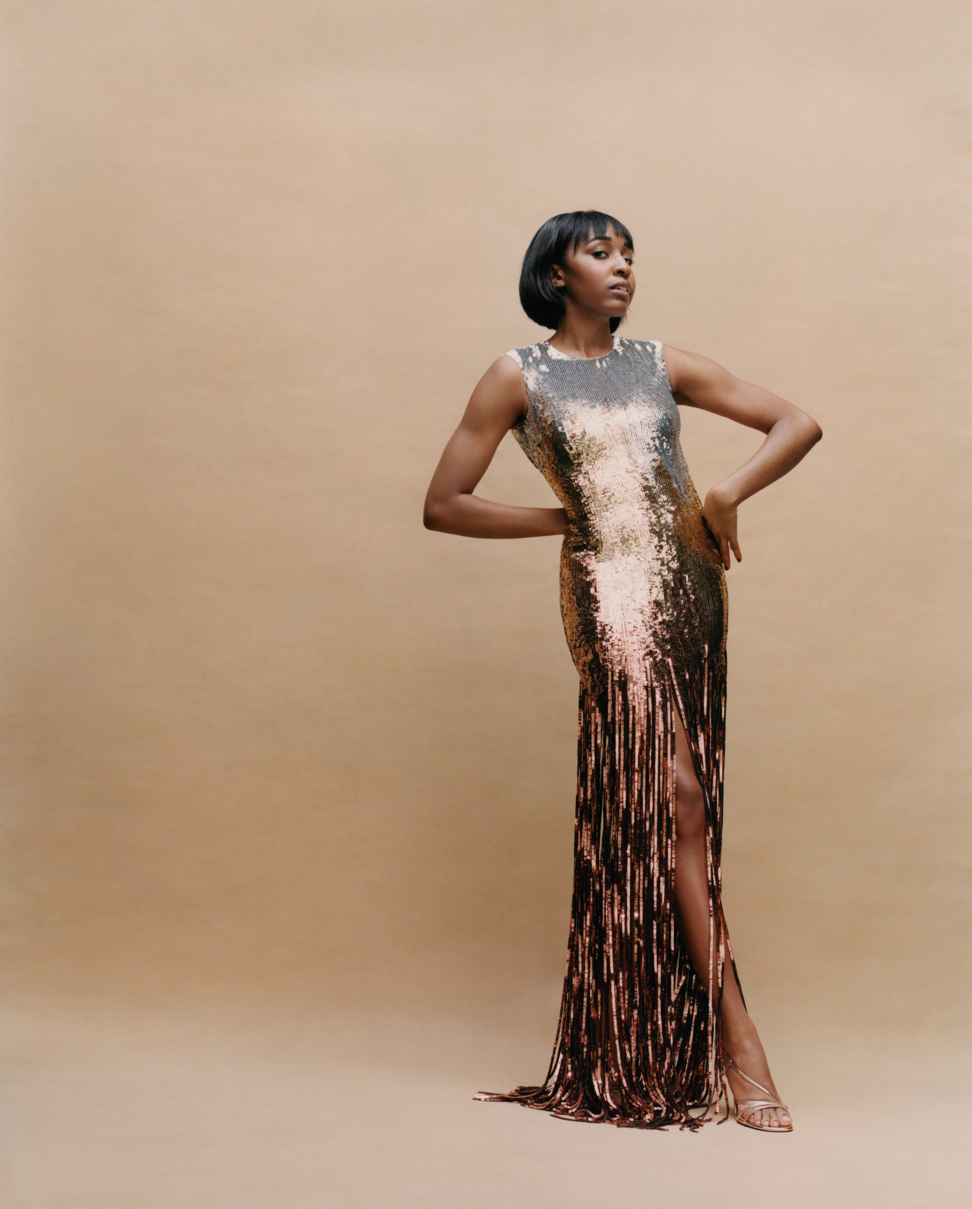 Ayo Edebiri posing in a sequined fringe gown against a neutral studio backdrop for Neiman Marcus.