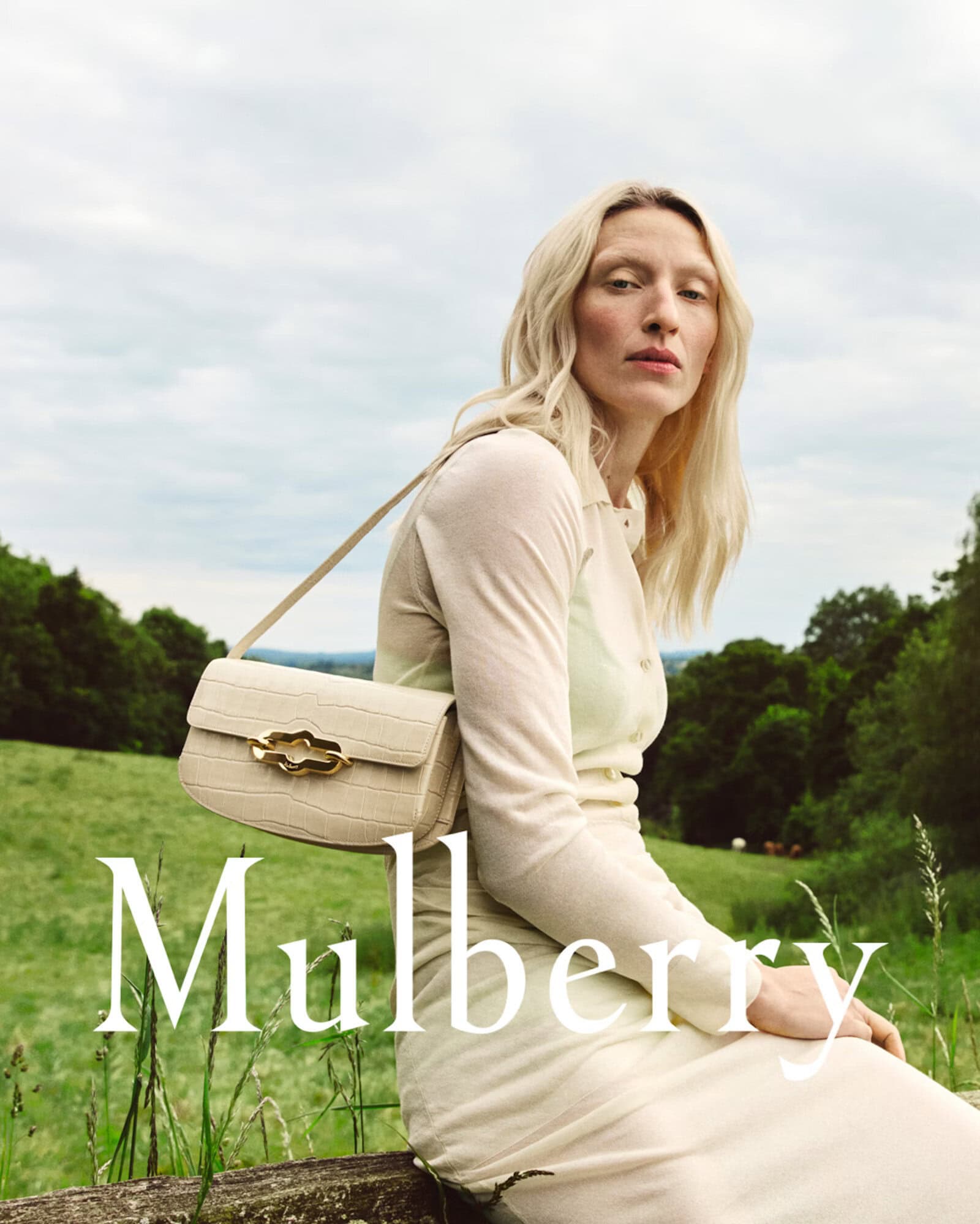 Blonde woman wearing cream knit dress holding textured shoulder bag in lush green countryside