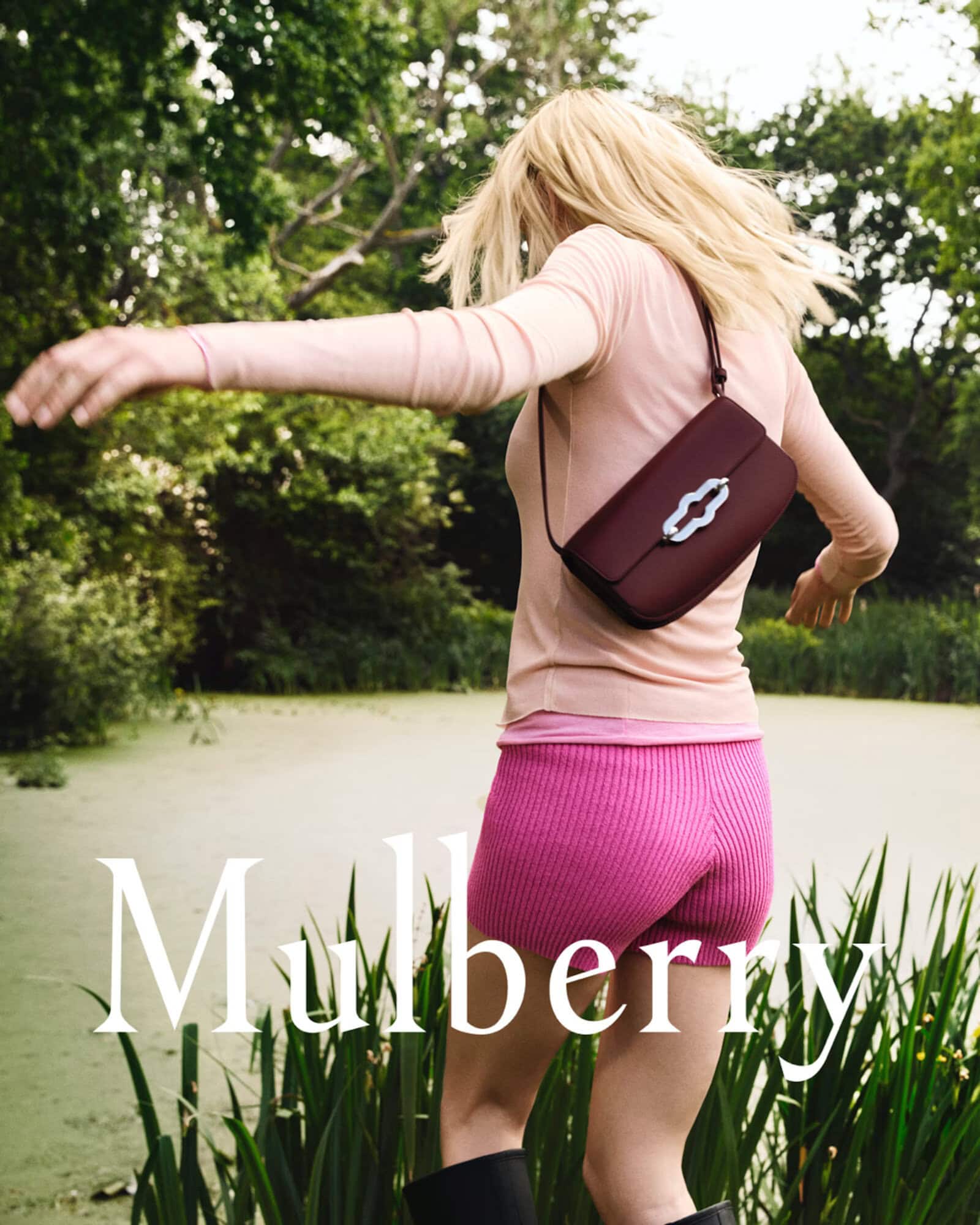Model with blonde hair spinning outdoors by a reedy pond wearing a pink top and shorts with a dark red shoulder bag.