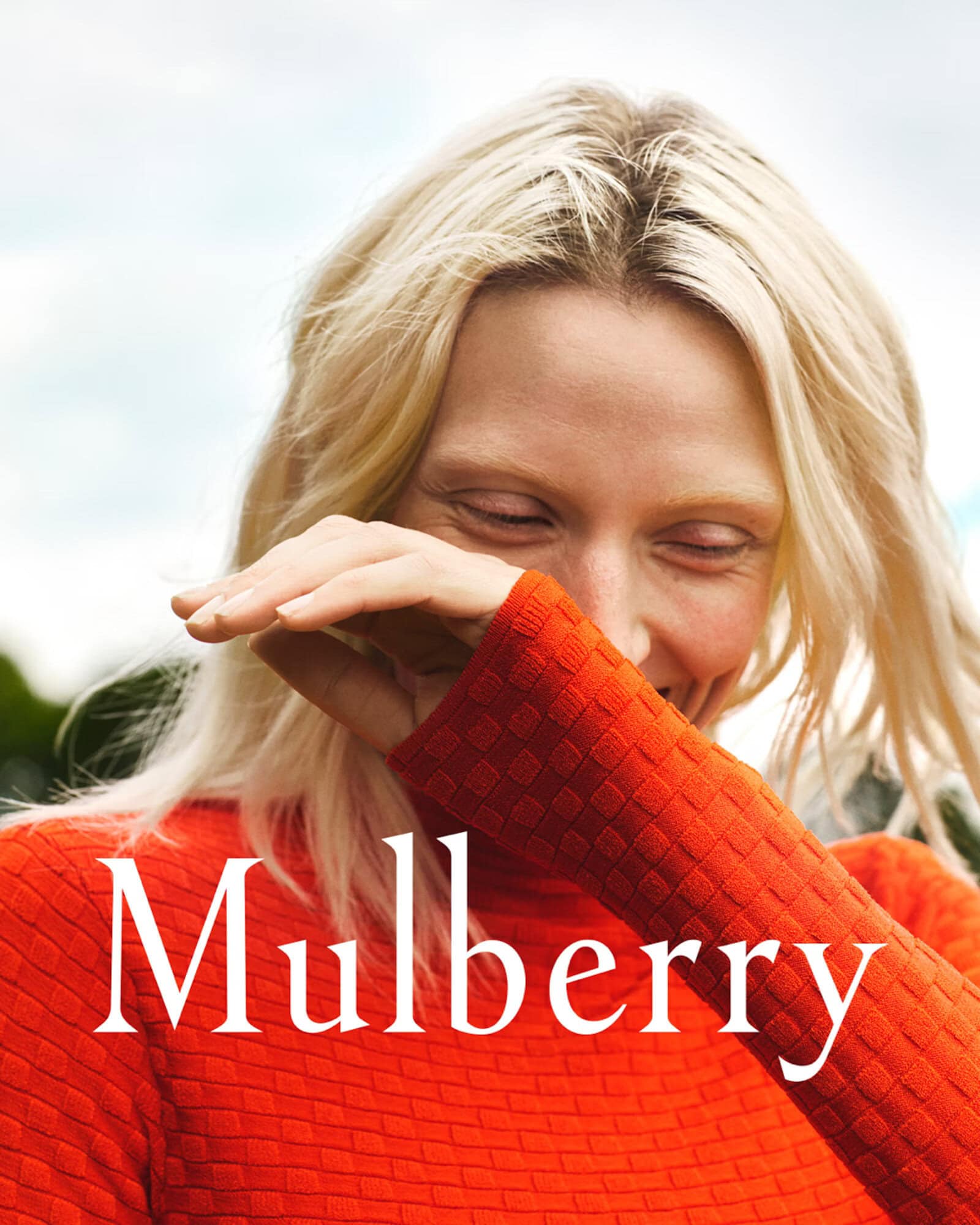Close-up portrait of a blonde woman wearing a textured orange sweater, shielding her eyes with her hand, against an outdoor background.