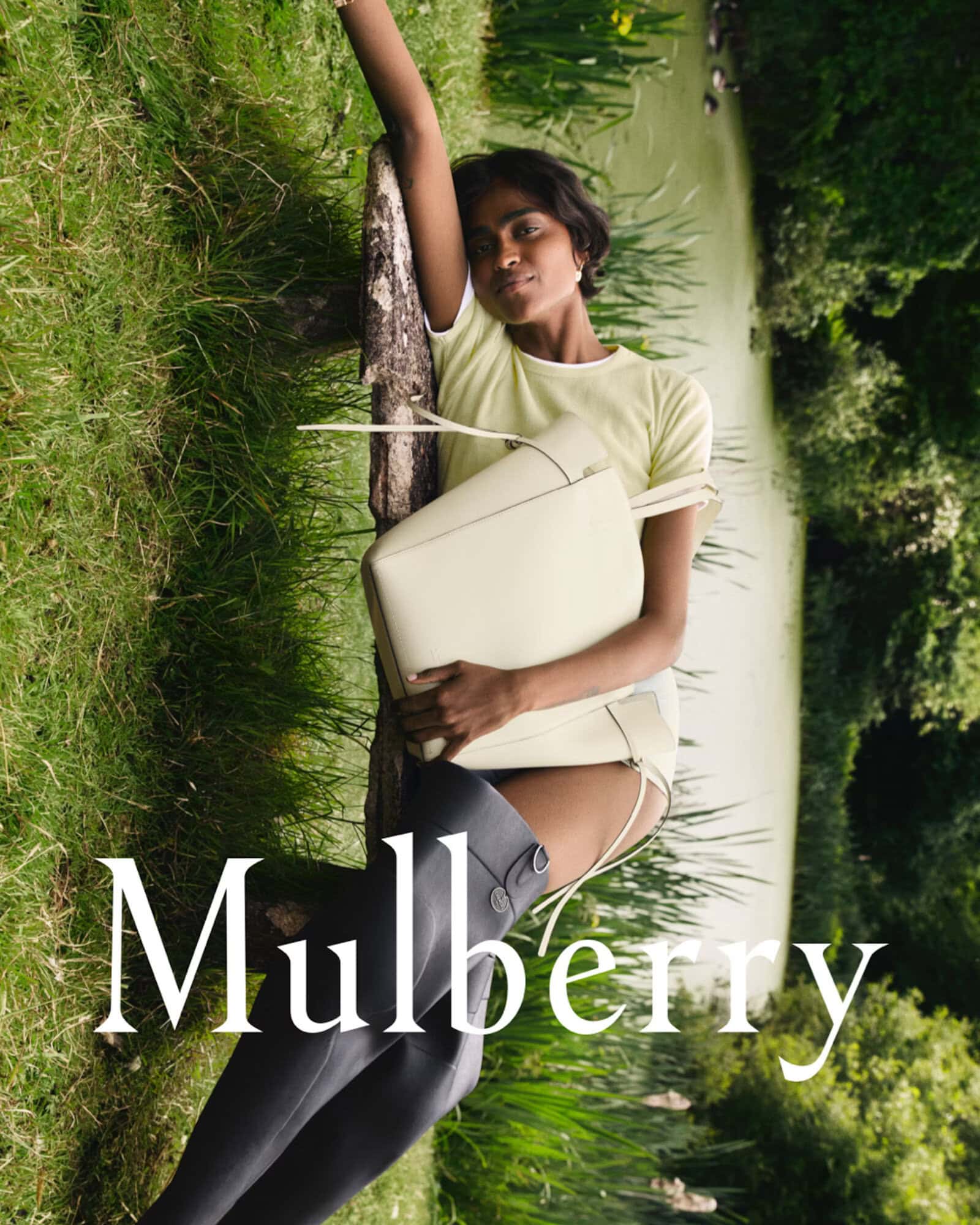 Model reclining against a wooden log holding a cream leather tote bag beside a grassy riverbank in a campaign by Guen Fiore for Mulberry.