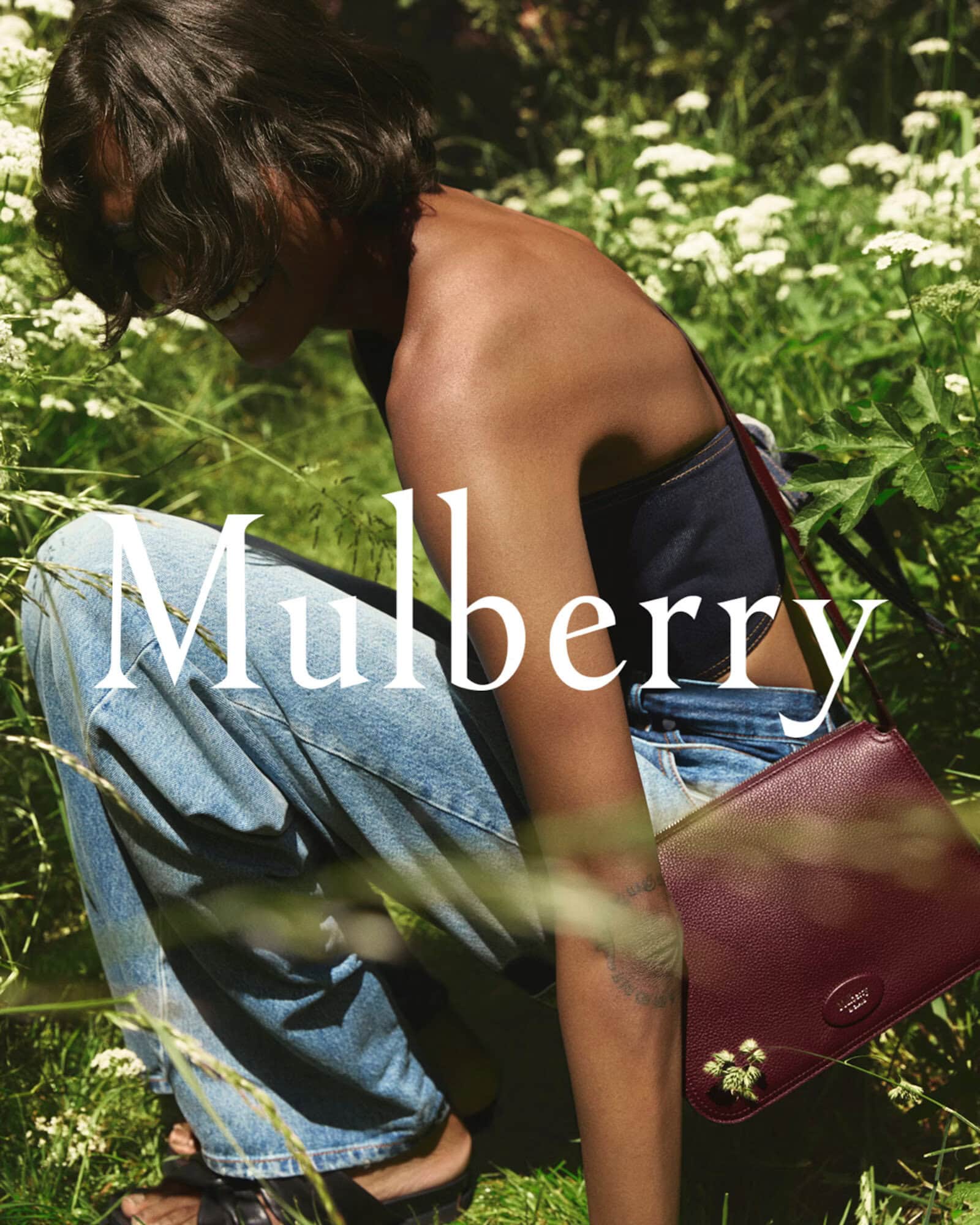 Model crouching outdoors in tall green grass wearing denim jeans and a burgundy leather shoulder bag for Mulberry