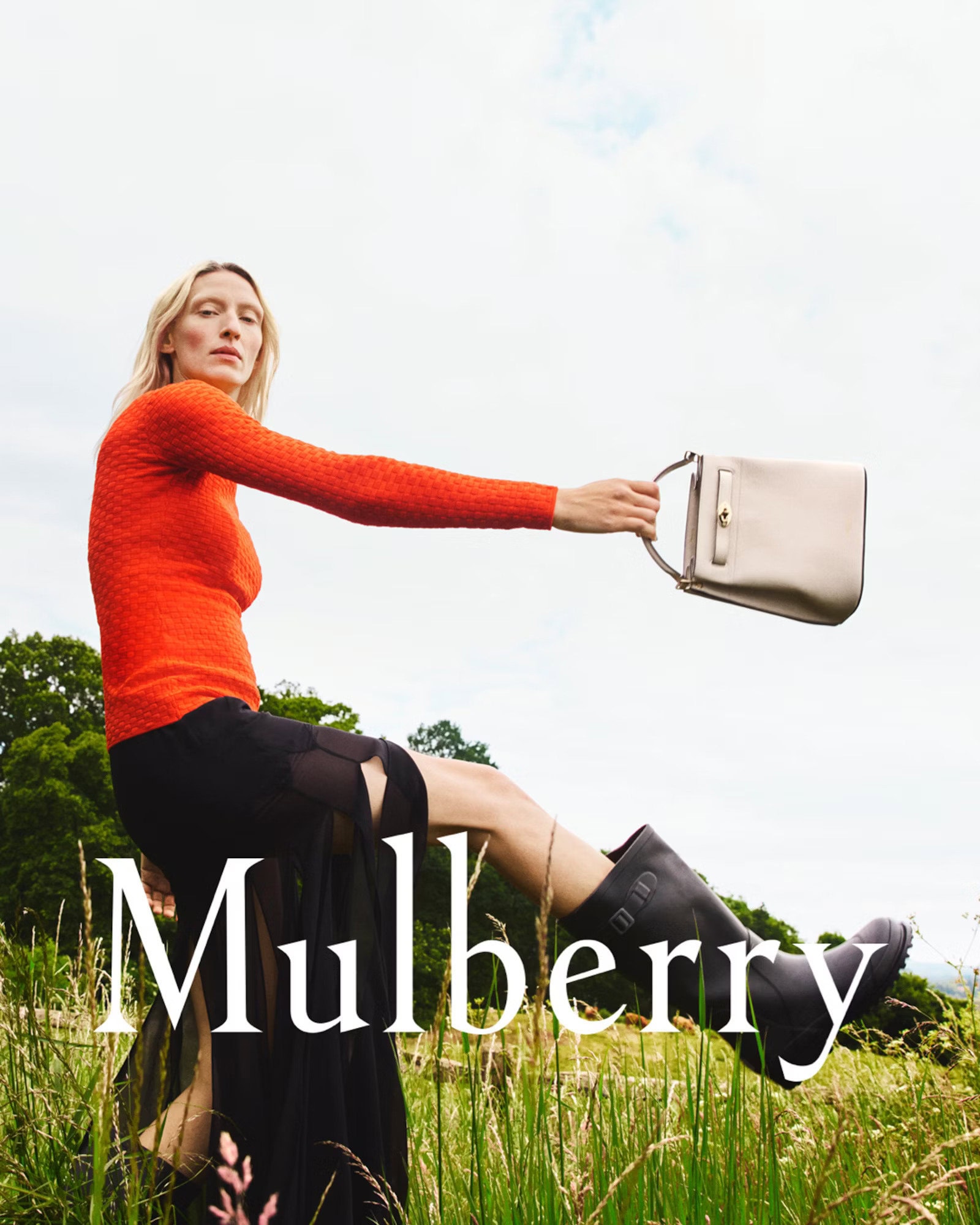 Model in orange sweater holding a handbag outdoors against a sky and meadow background