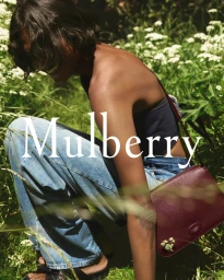 Model crouching outdoors in tall green grass wearing denim jeans and a burgundy leather shoulder bag for Mulberry