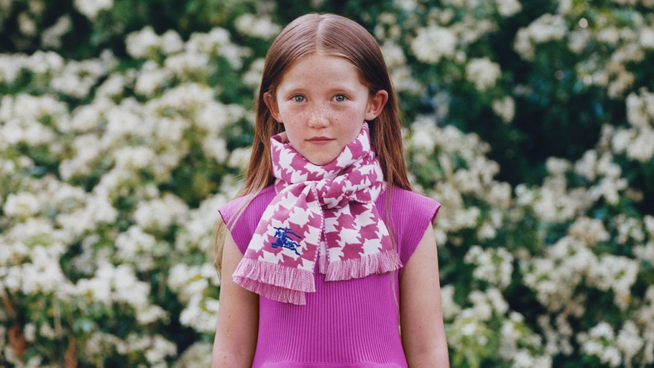 Young child wearing a pink knit dress and patterned scarf outdoors against a background of white flowering bushes