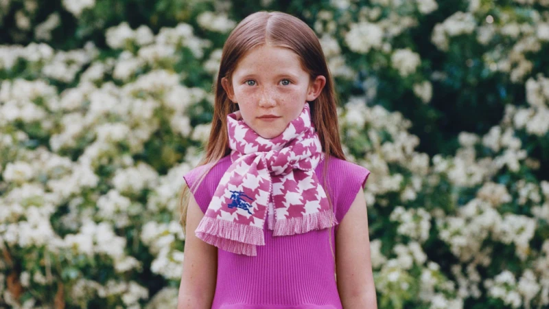 Young child wearing a pink knit dress and patterned scarf outdoors against a background of white flowering bushes