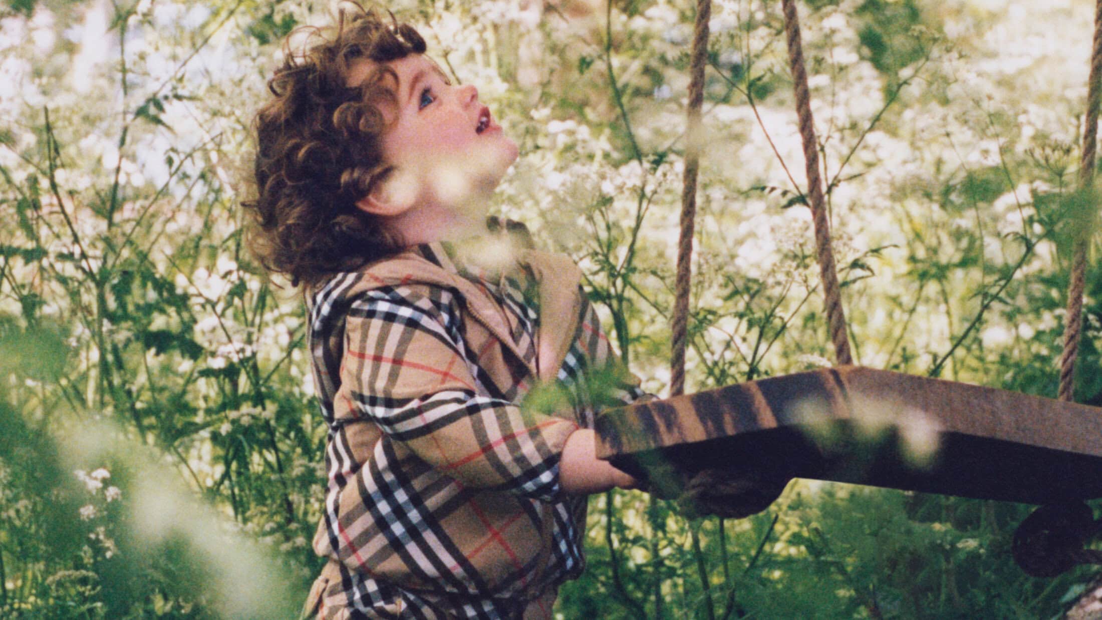 A young child wearing a plaid jacket holding onto rope swings in a lush outdoor setting