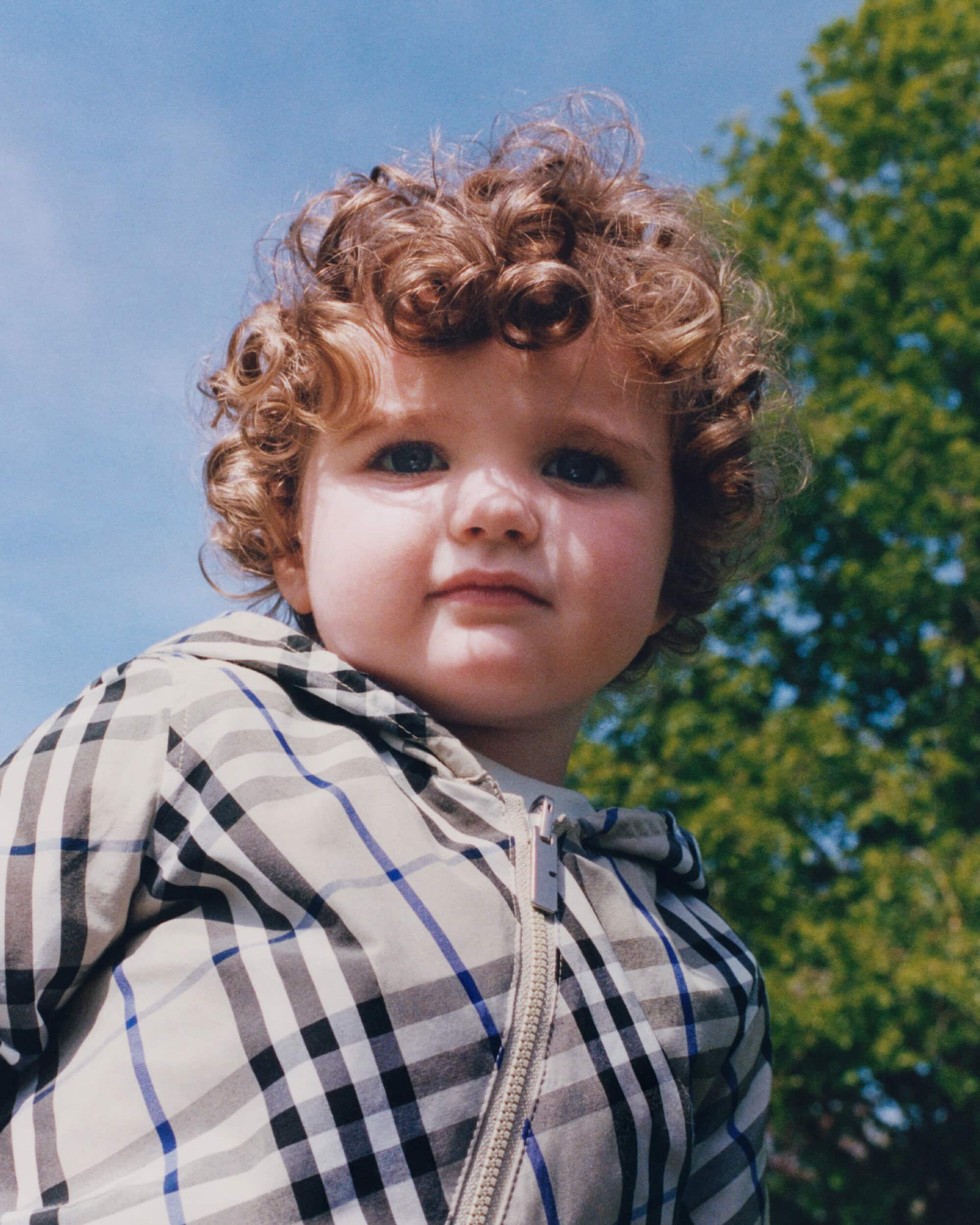 Young child with curly hair wearing a plaid hooded jacket outdoors against a clear blue sky and green trees.