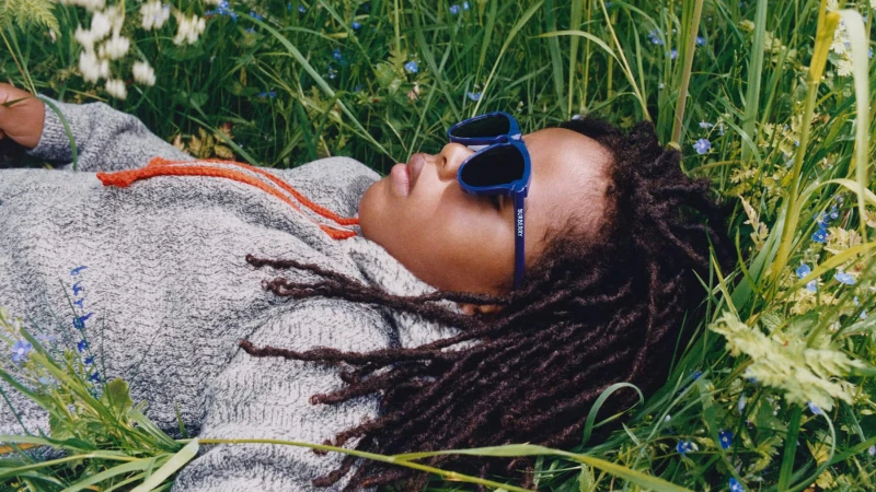 Child with dark locs wearing blue sunglasses lying in tall green grass for Burberry winter campaign