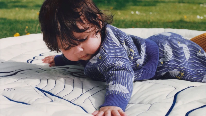 Baby wearing a patterned blue knit sweater lying on a quilted blanket outdoors in a grassy park