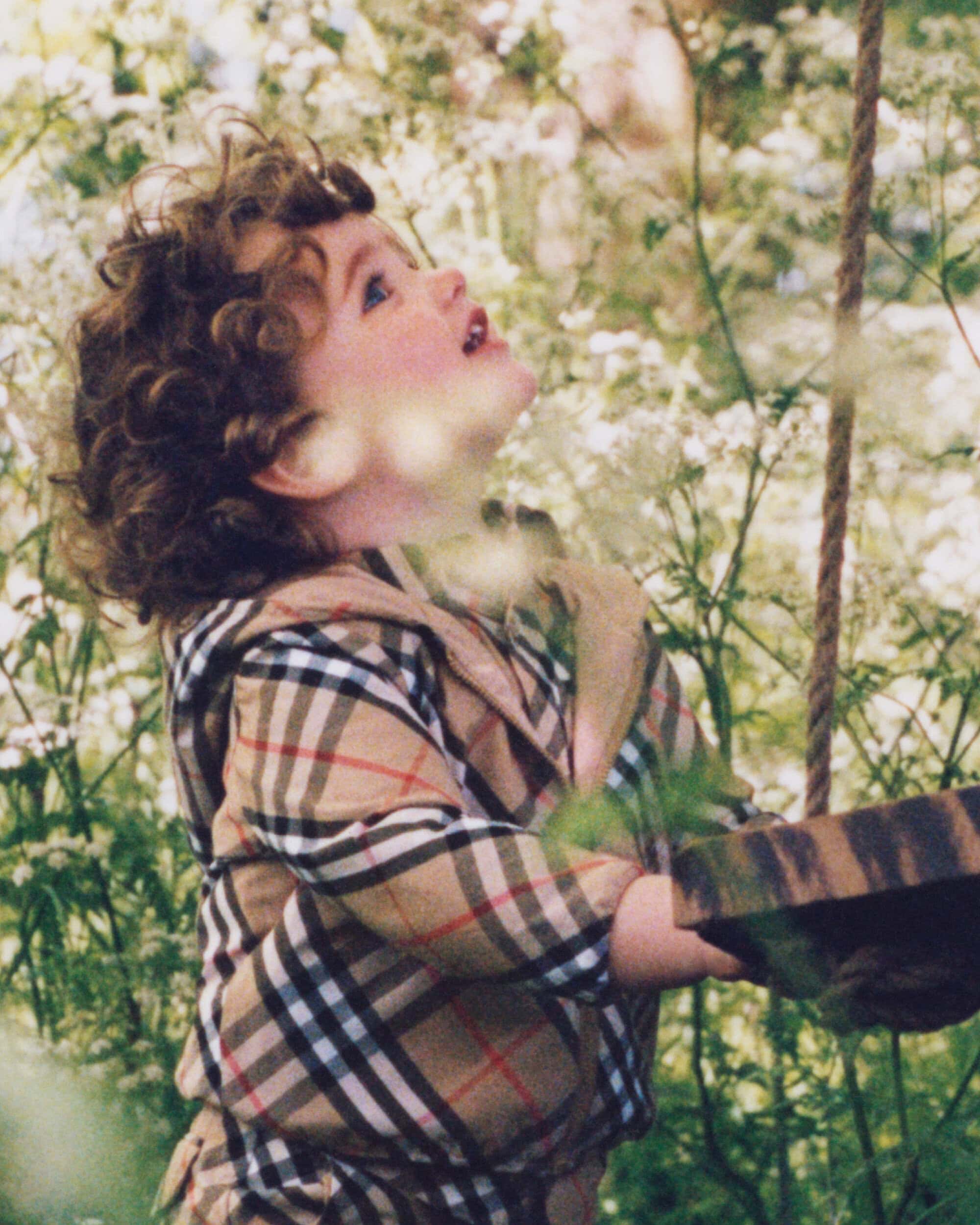 A young child with curly hair wearing a plaid jacket looking up while standing outdoors near foliage.