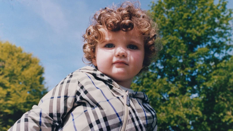 Child with curly hair wearing a plaid zip-up jacket outdoors against green trees and blue sky