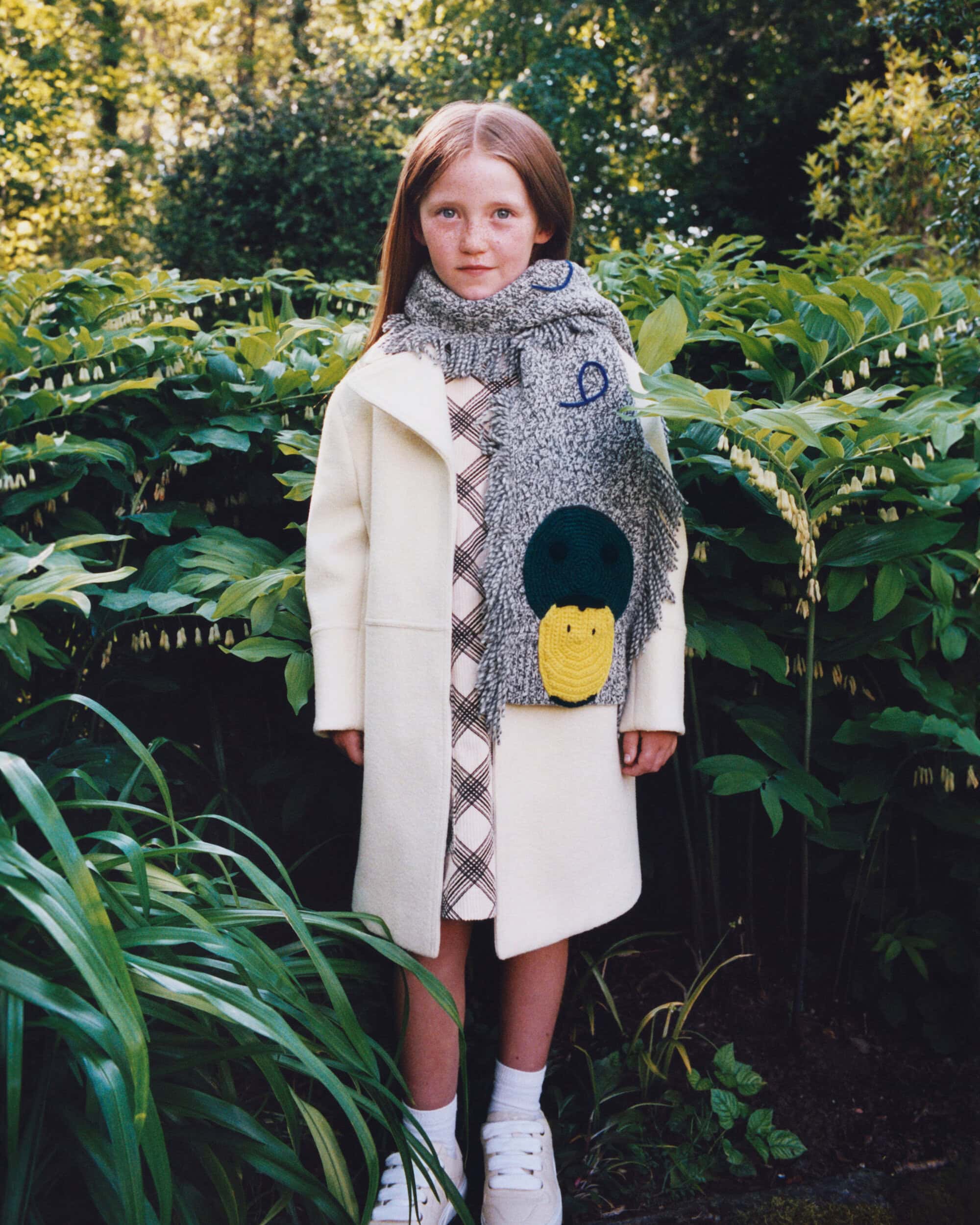 Young girl wearing a cream coat and duck scarf standing outdoors among lush green foliage for Burberry.