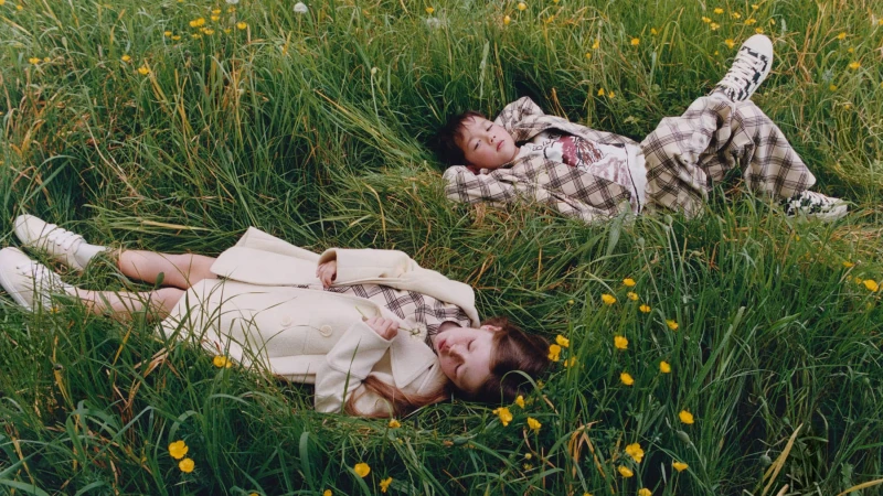 Two children lying down in a tall green meadow filled with yellow wildflowers, wearing plaid clothing.