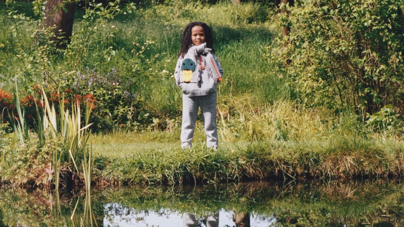 A child in a grey patterned sweater and matching trousers stands near a grassy bank reflecting in water.