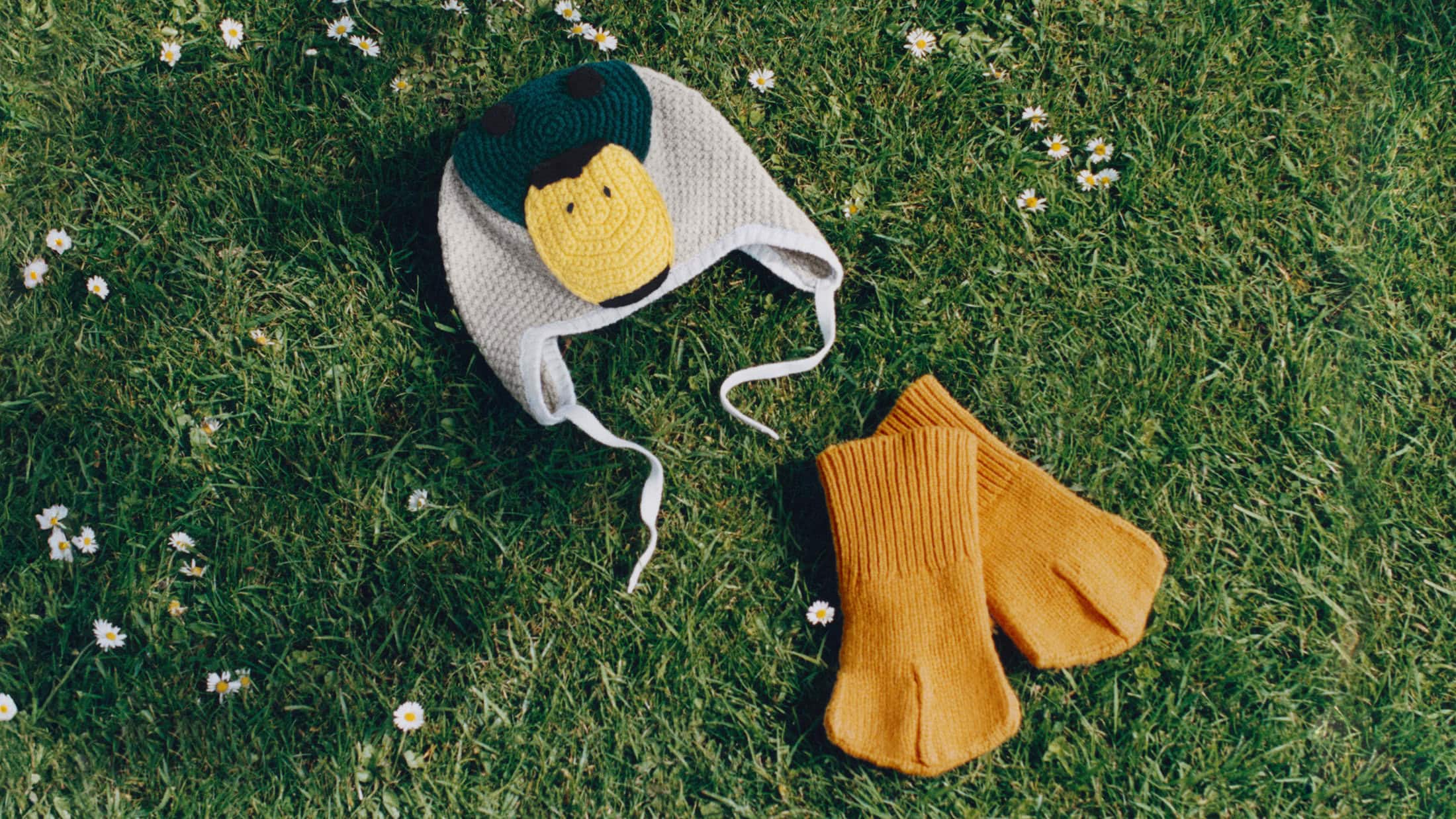 Knit duck cap and yellow mittens resting on a green grass lawn dotted with small white daisies.