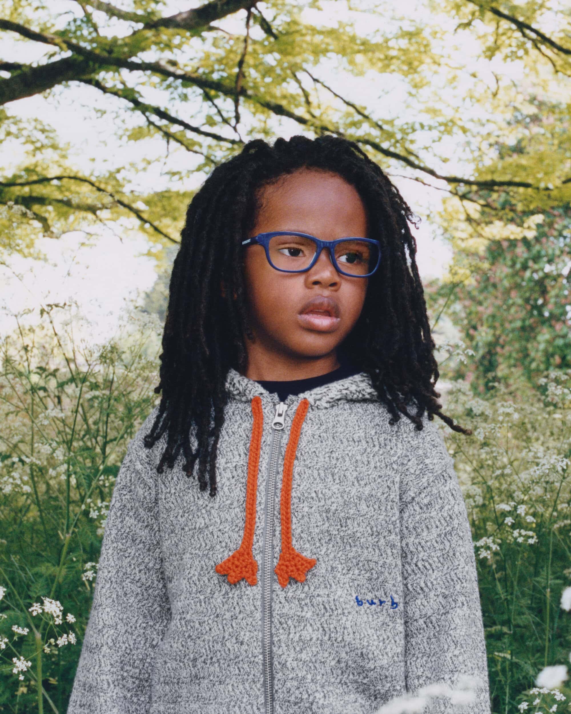 Child with locs wearing a grey zip hoodie and blue glasses standing among wildflowers outdoors