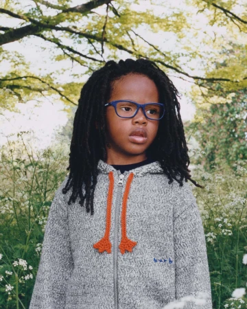 Child with locs wearing a grey zip hoodie and blue glasses standing among wildflowers outdoors