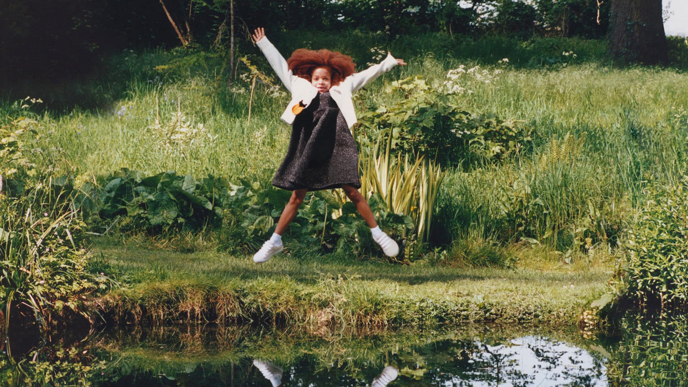 A young child jumping mid-air in a lush green meadow near a water edge for the Burberry campaign.