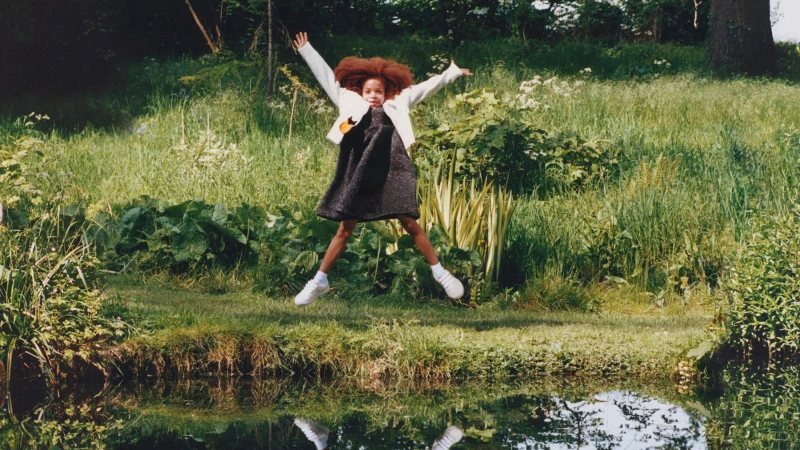 A young child jumping mid-air in a lush green meadow near a water edge for the Burberry campaign.