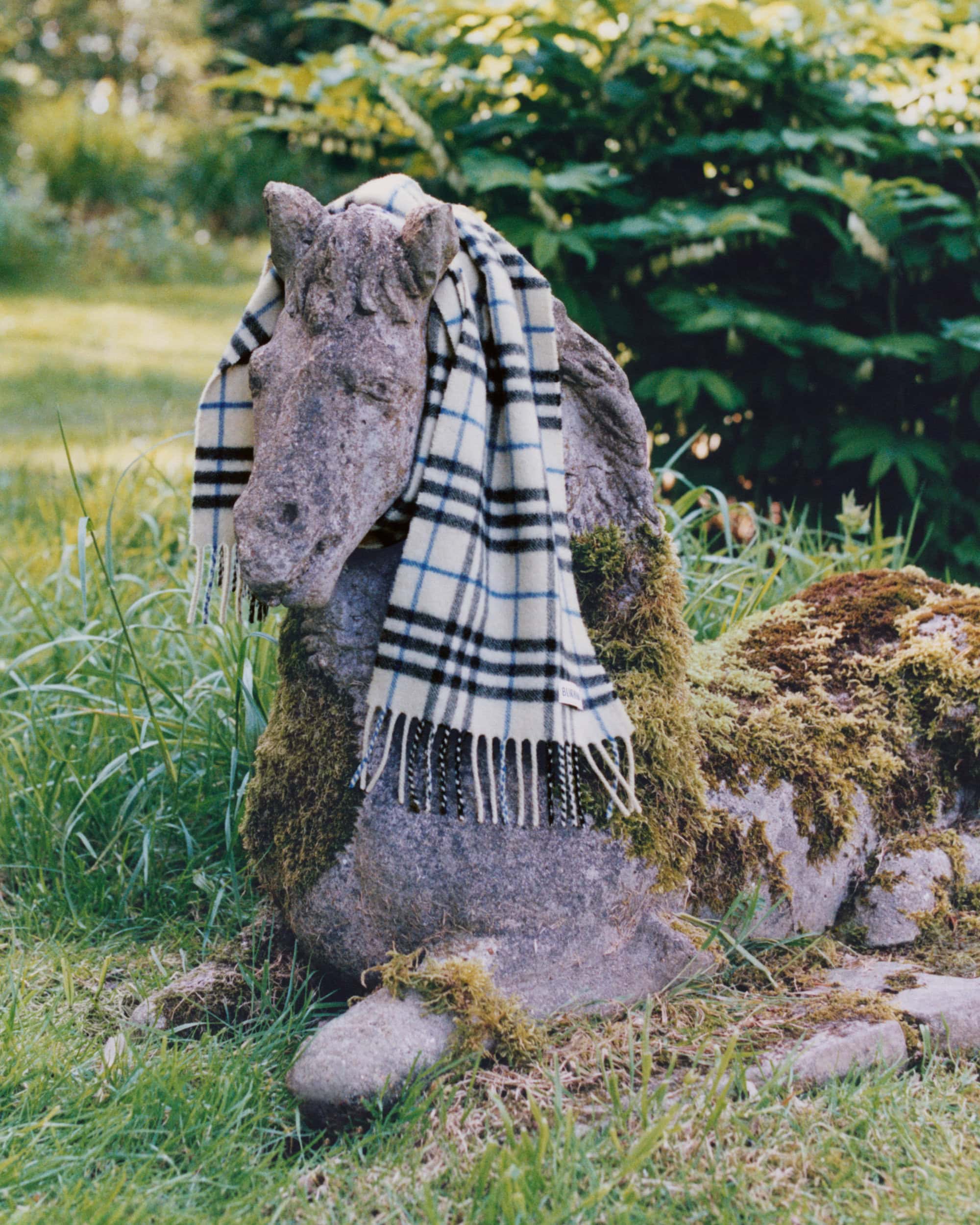 Stone horse statue covered in moss wearing a plaid patterned scarf outdoors in a grassy garden setting.
