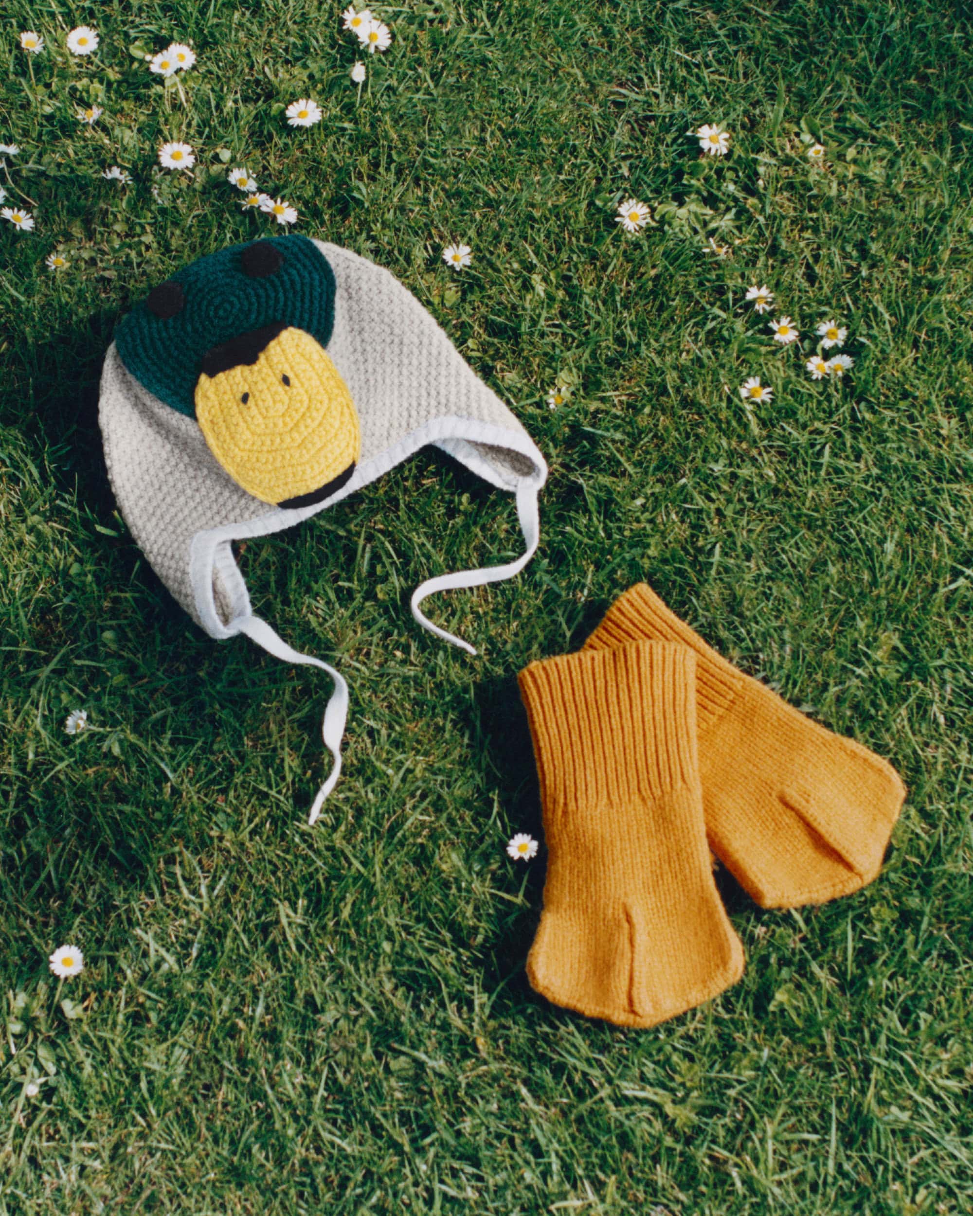 Knit duck bonnet and yellow webbed mittens laid out on green grass with scattered small white flowers.