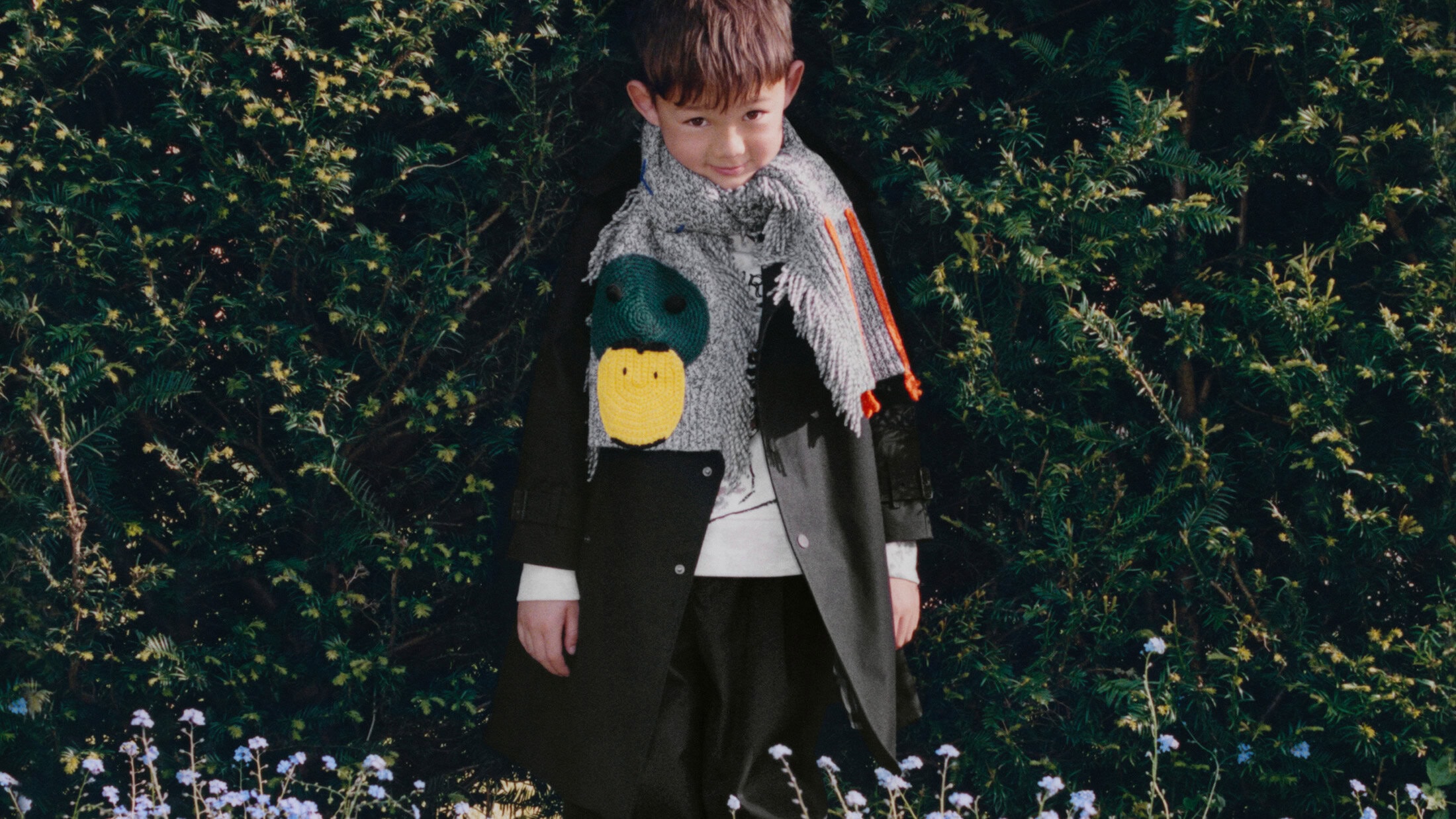 Child wearing a dark coat and a grey fringed scarf with a duck motif outdoors against green foliage for Burberry winter campaign
