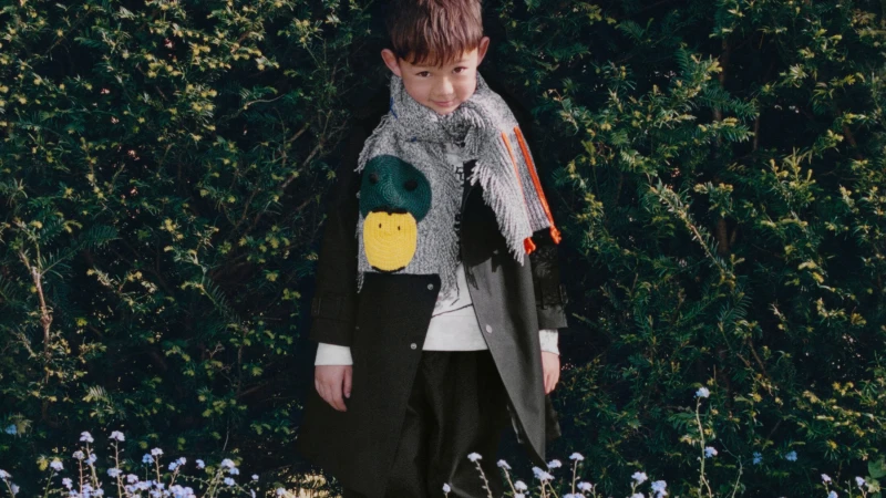 Child wearing a dark coat and a grey fringed scarf with a duck motif outdoors against green foliage for Burberry winter campaign
