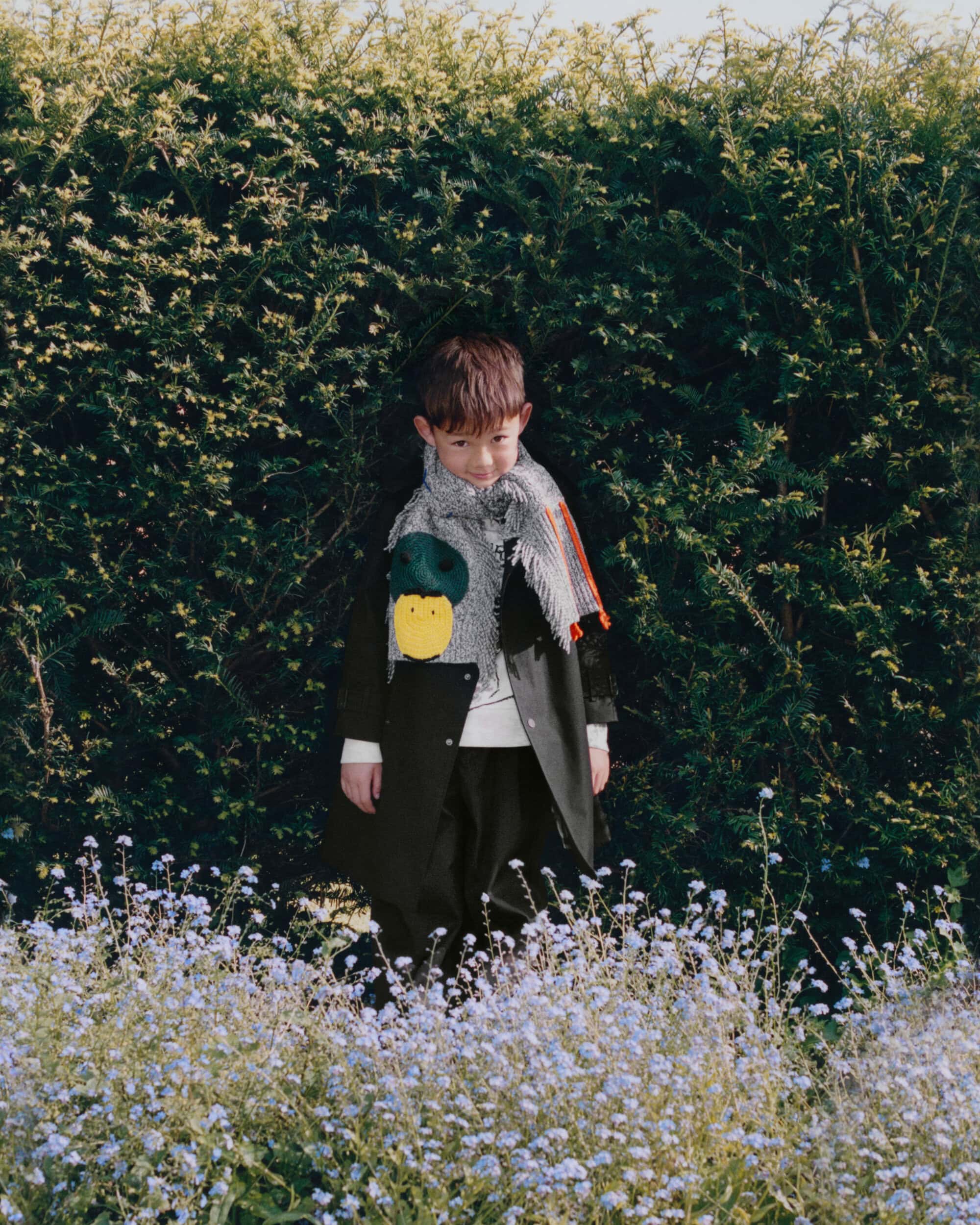 Young child standing in front of a dense green hedge wearing a dark coat and fringed scarf with playful patches