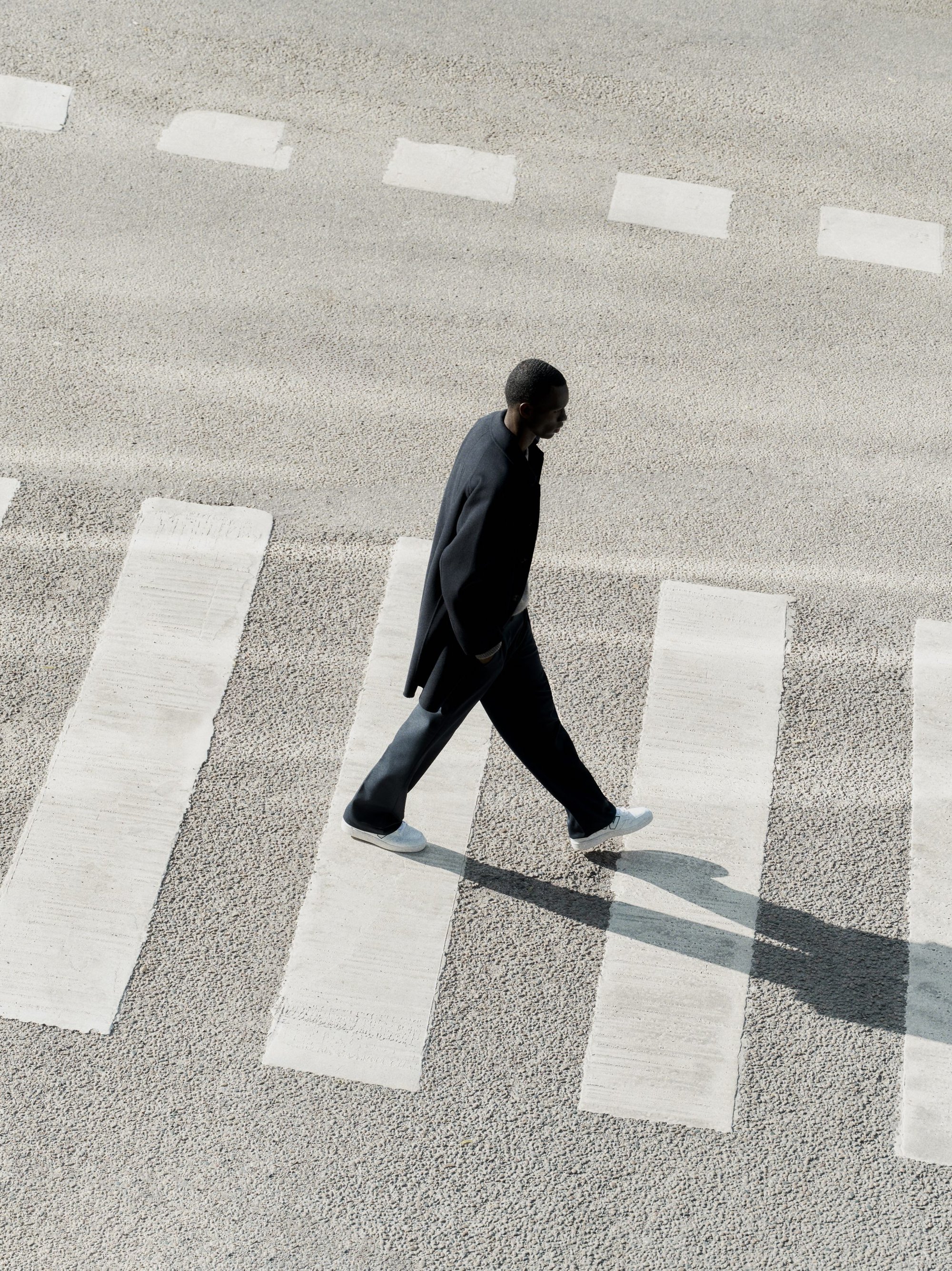 High-angle view of a man walking across a striped pedestrian crosswalk wearing a dark coat and light sneakers.