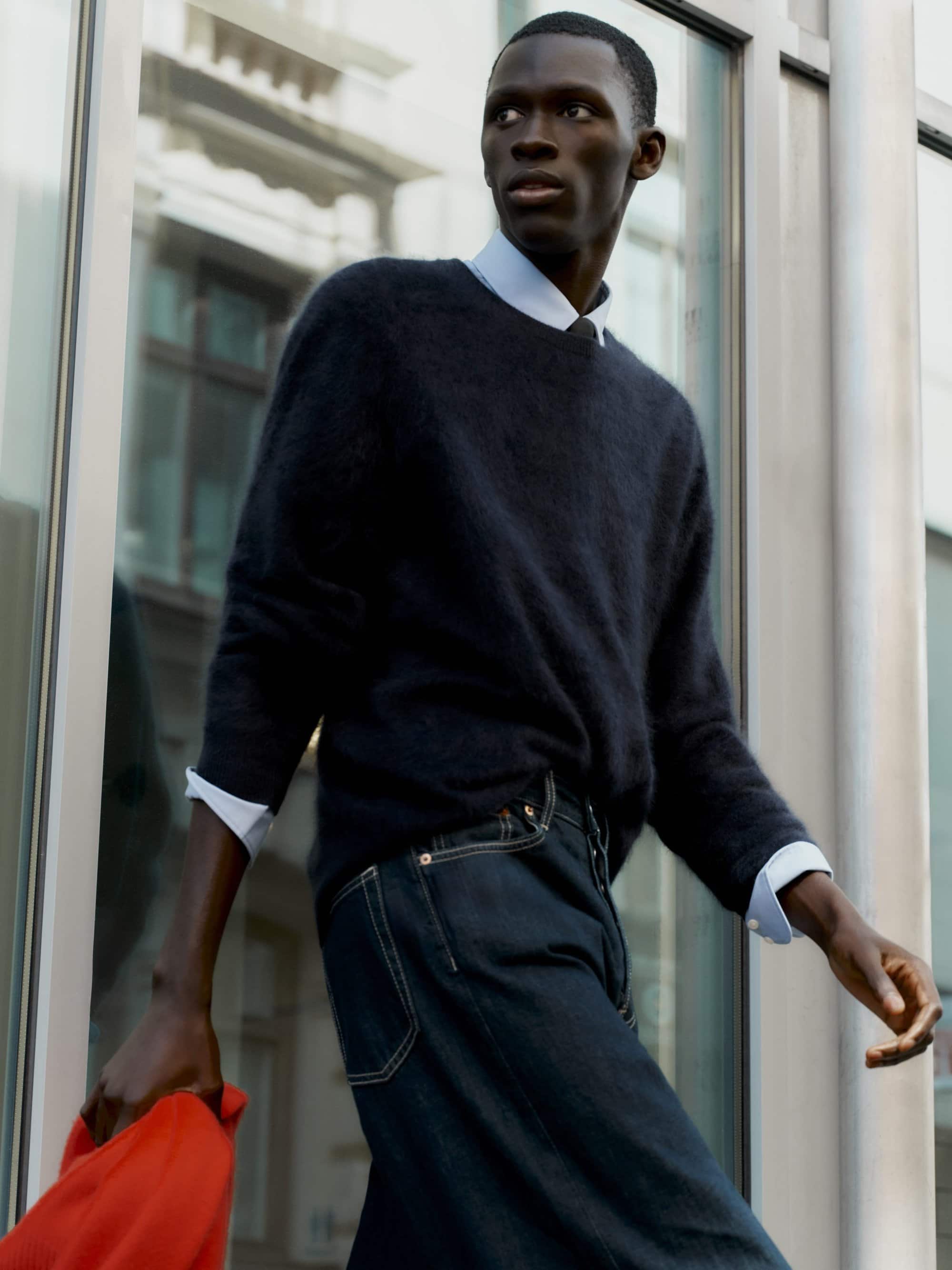 Black man in a dark knit sweater and blue jeans walking past a building exterior, holding a red garment.