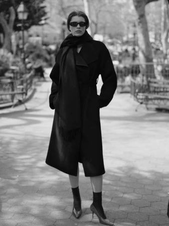 Model in dark sunglasses wearing a long black coat and scarf standing on a paved path