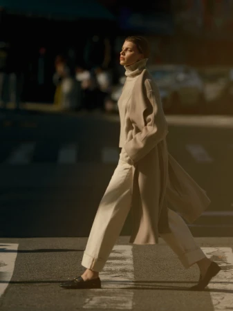 Model wearing a beige coat and trousers walking across a city street during the day