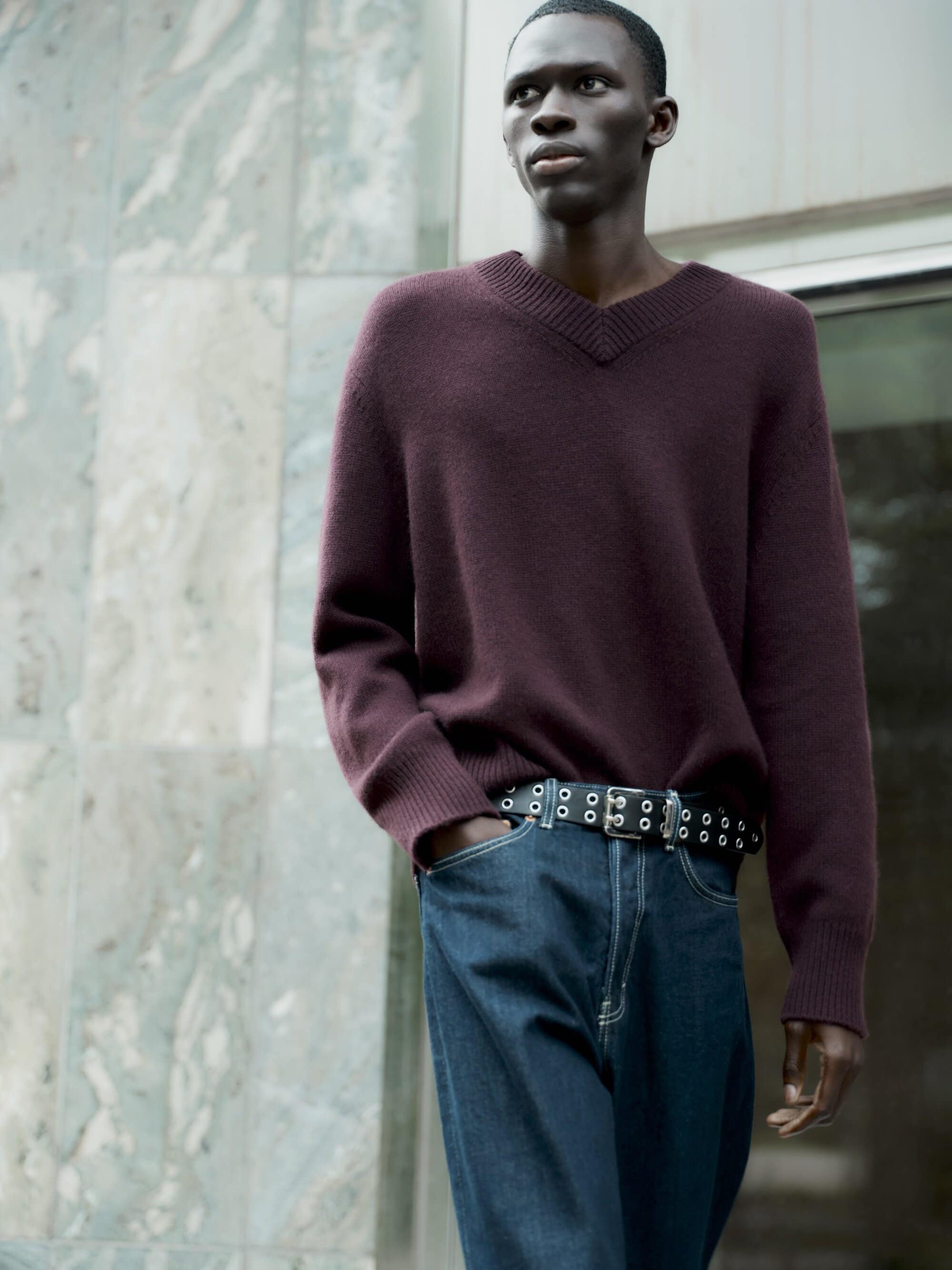 Man wearing burgundy V-neck sweater and blue jeans with grommet belt outdoors against stone building facade