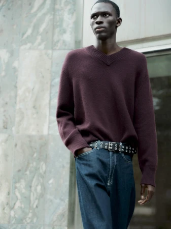Man wearing burgundy V-neck sweater and blue jeans with grommet belt outdoors against stone building facade