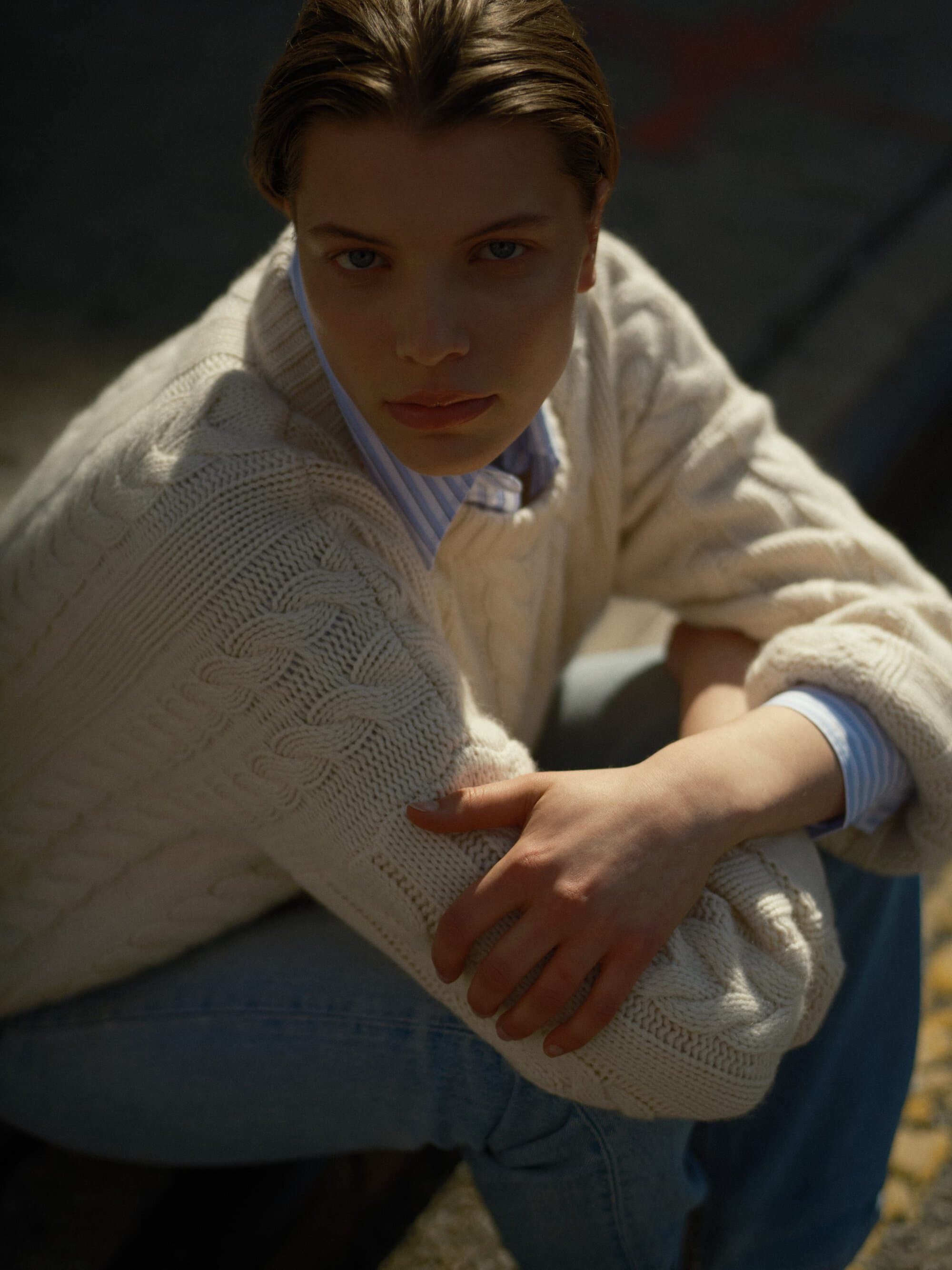 Woman wearing a cream cable-knit sweater and blue jeans posing outdoors for Soft Goat Fall 2024 ad campaign.