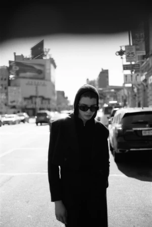 Model in dark sunglasses and hooded jacket walking on an urban street with cars and buildings in the background.